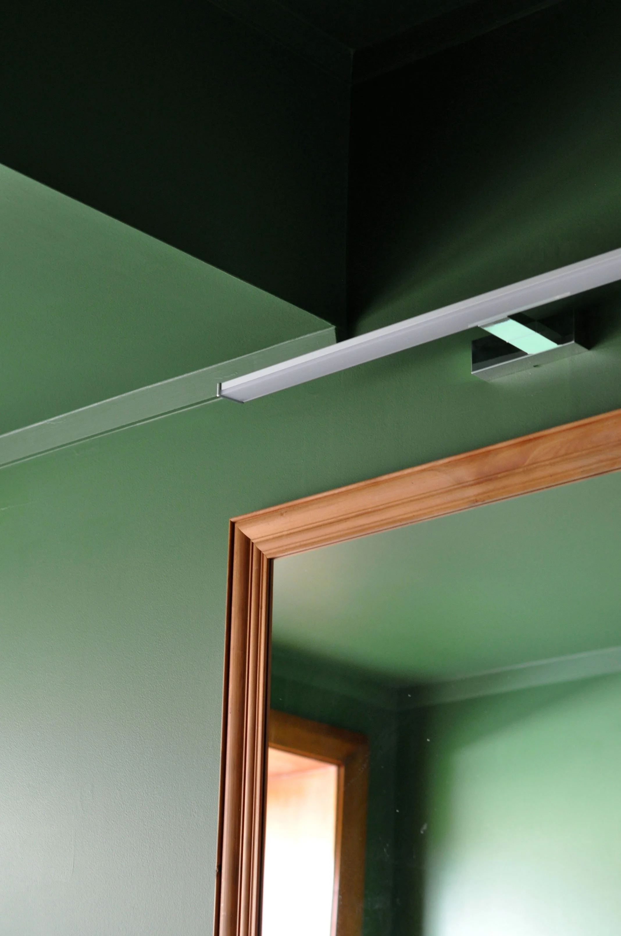 The image shows a reflection of an open doorway with a wooden frame in a mirror. The wall around the mirror is painted green, and there is a ceiling with a fluorescent light fixture.