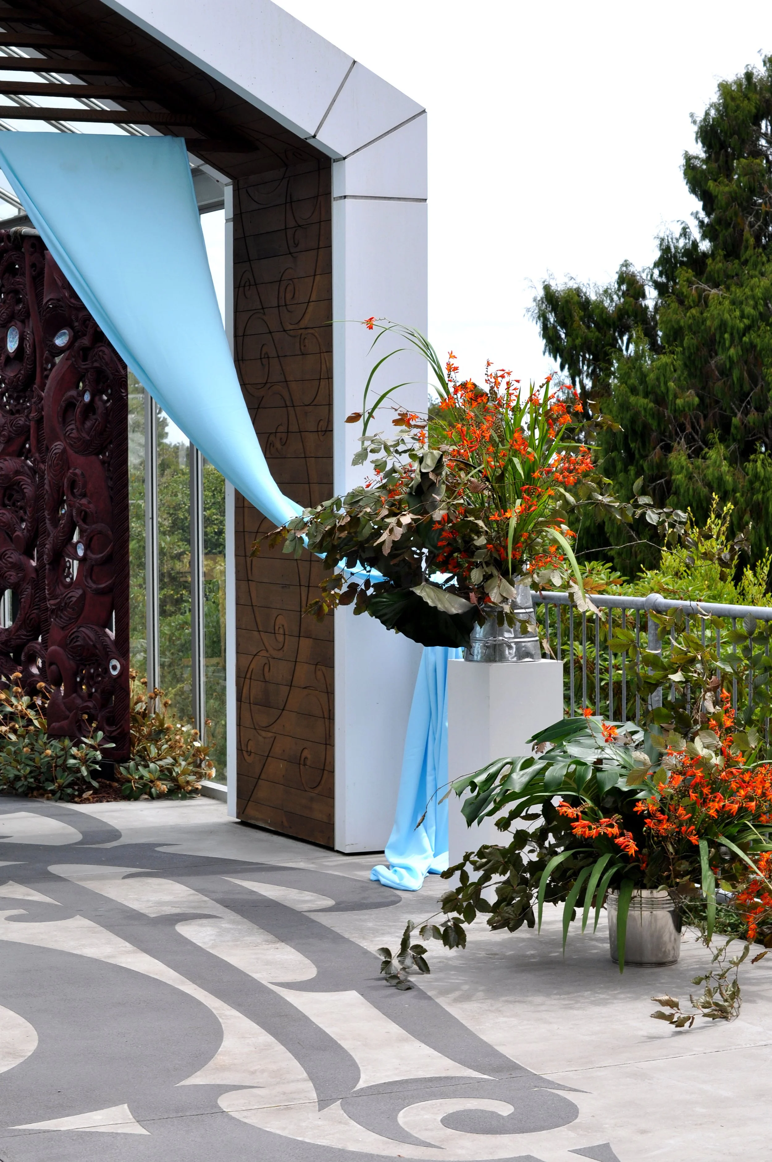 A decorative outdoor space with a large vase of orange flowers, surrounded by various green plants. Part of a modern architectural structure with wood paneling, white walls, and a metal railing, with trees in the background.