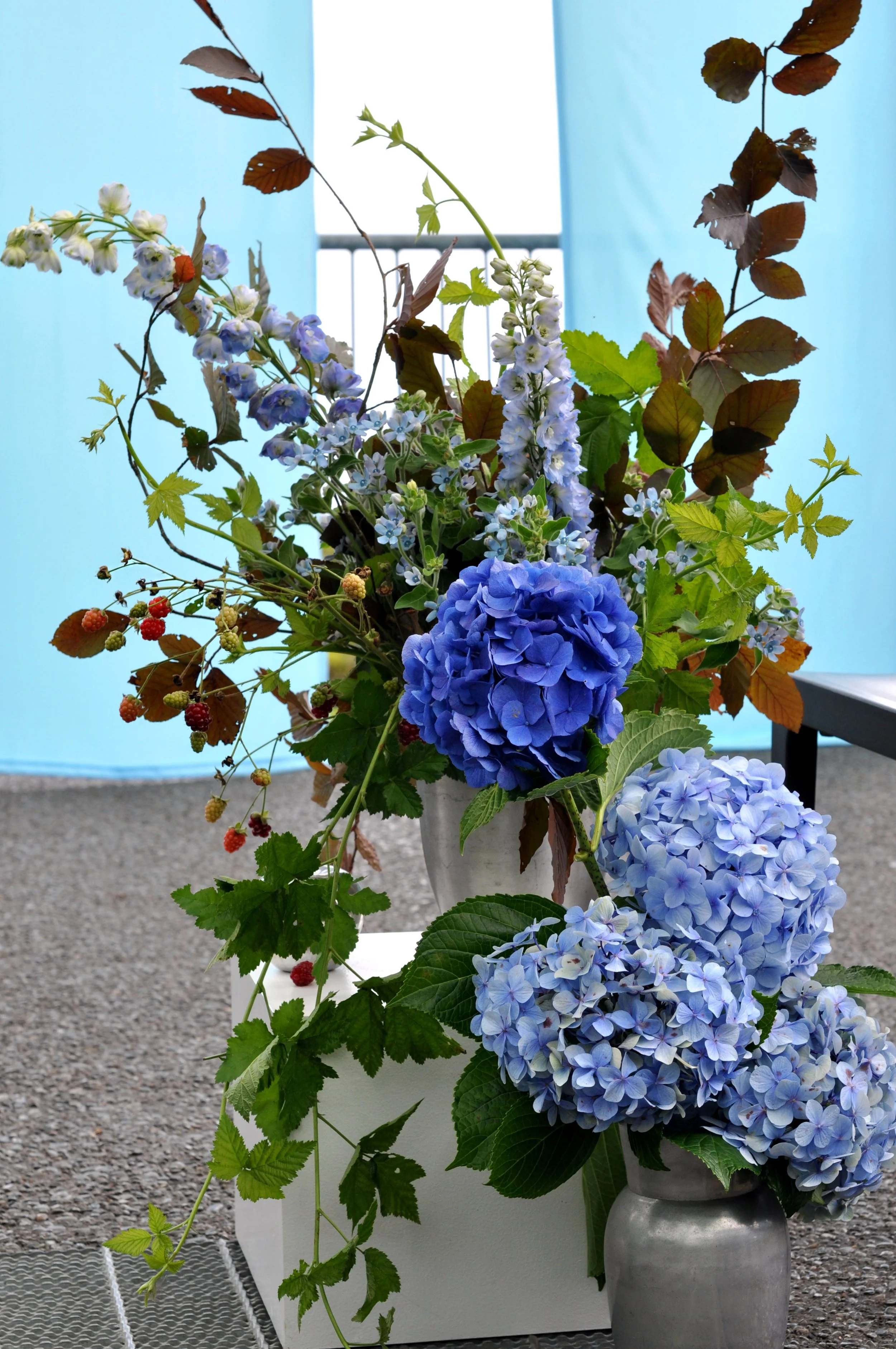 A bouquet of various blue, purple, and white flowers with green and brown leaves in a silver vase, placed on a textured gray surface against a background with light blue panels and a metal railing.