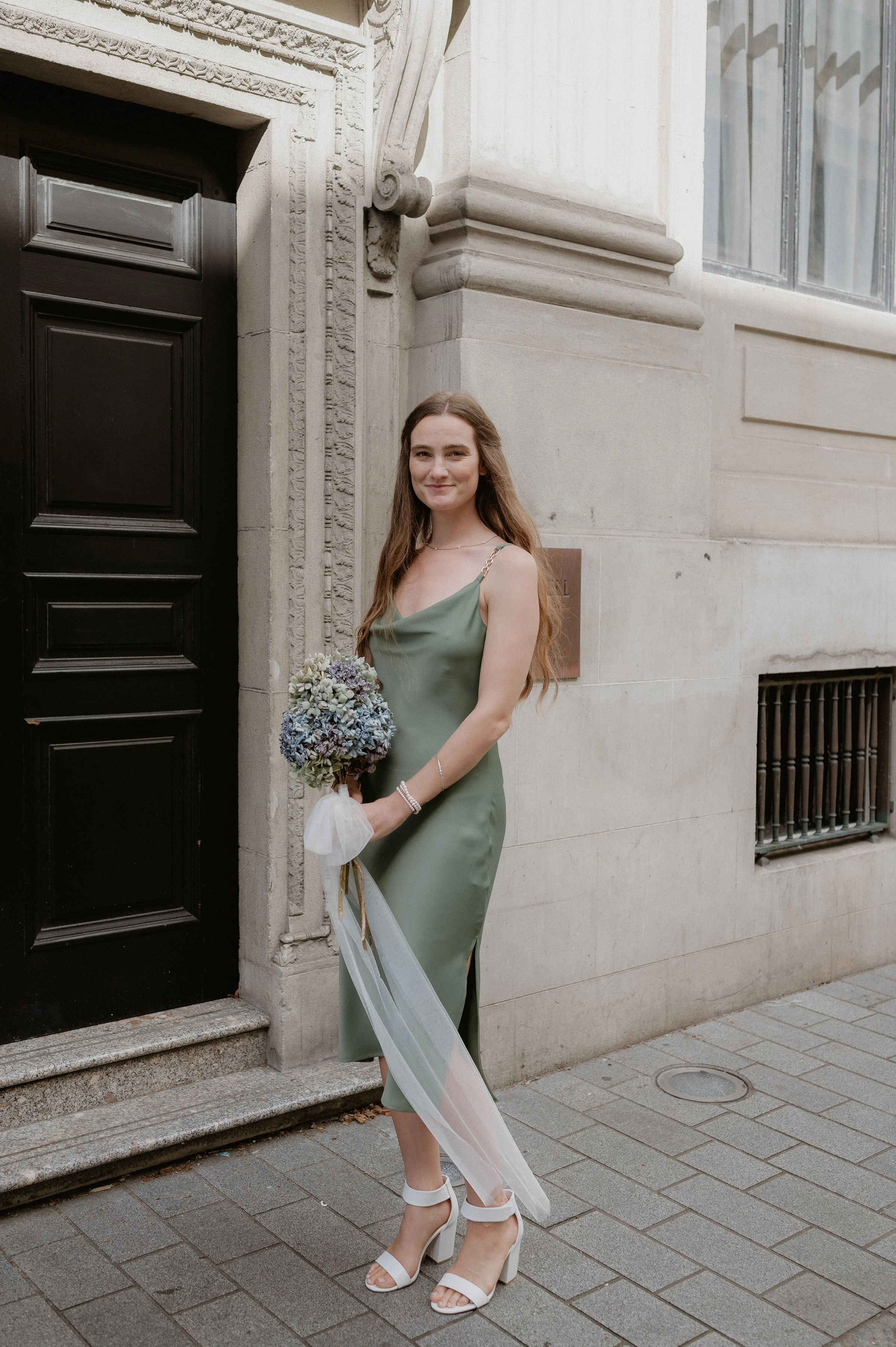 A young woman with long brown hair wearing a silk green dress and white high-heeled shoes holding a bouquet of flowers stands on a city sidewalk next to a building with stone architectural details and large windows.