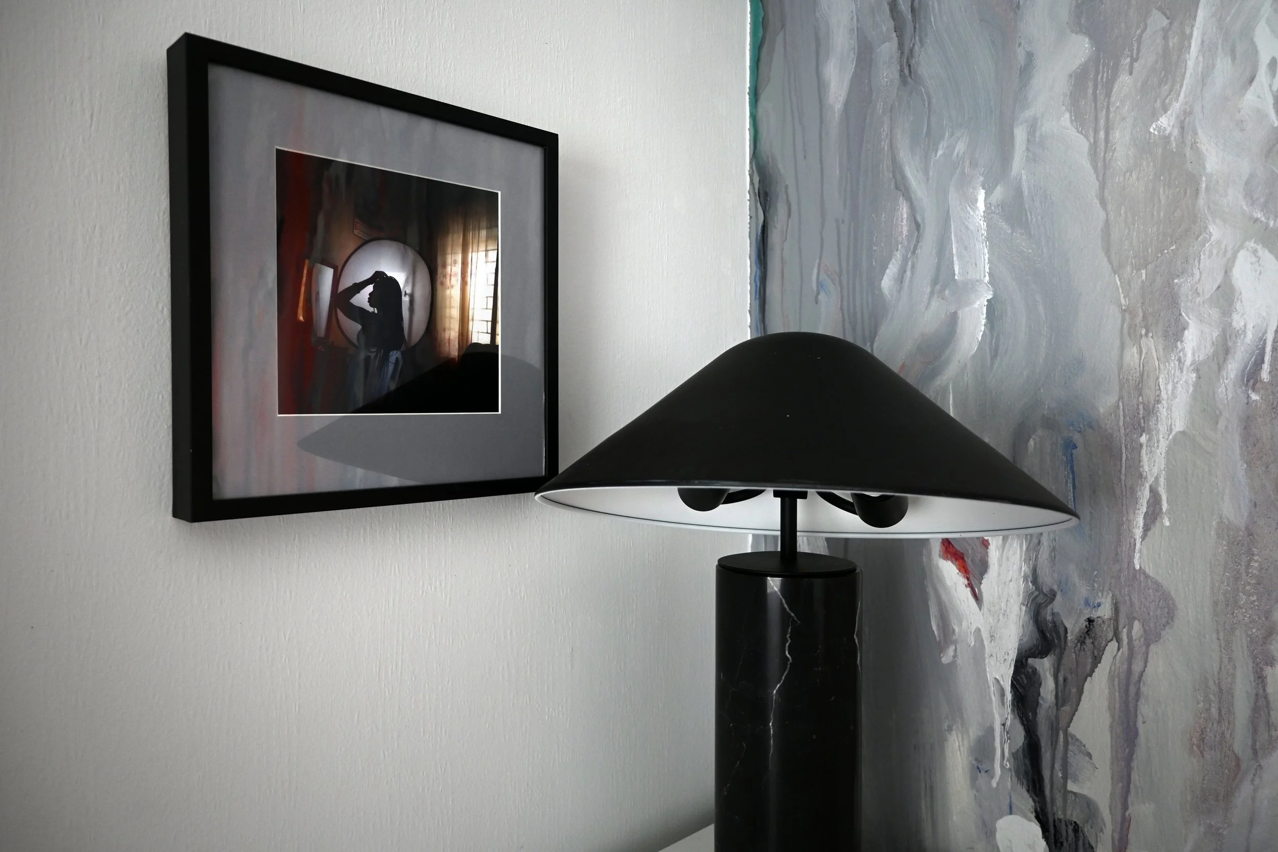 A framed photograph of a silhouette of a woman with long hair, hanging on a white textured wall, next to a table lamp on a black round surface with a marble pattern on the tabletop.