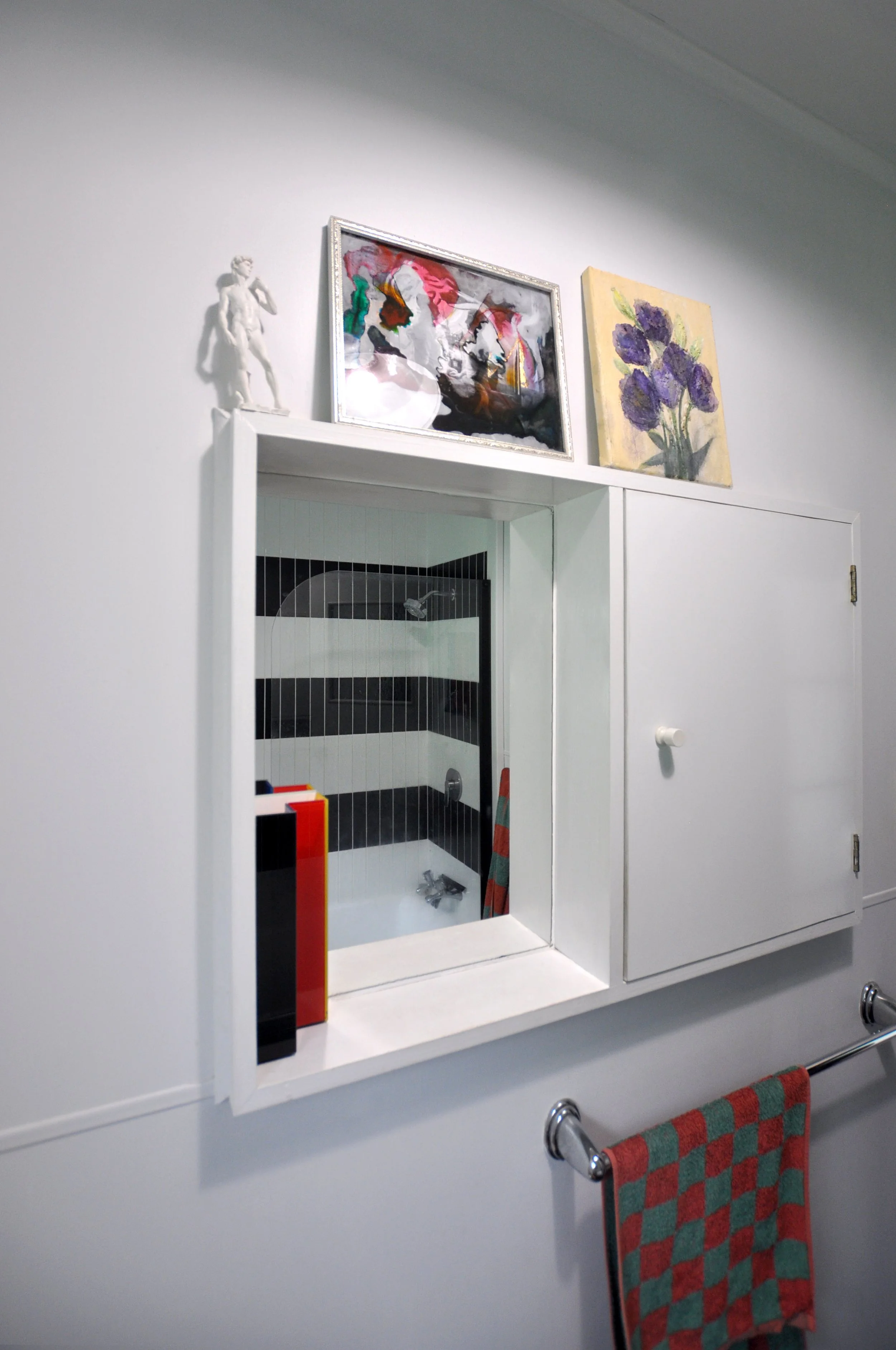 A white wall-mounted cabinet with a mirror, a small sculpture of a boy, and two framed paintings of flowers on top. A towel with a red and green checkered pattern hangs below.