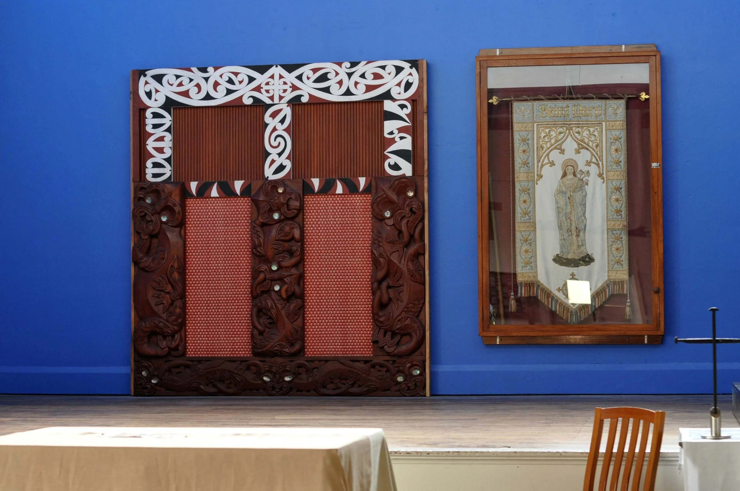 Cultural display in museum with carved wooden panel on blue wall next to framed religious tapestry.
