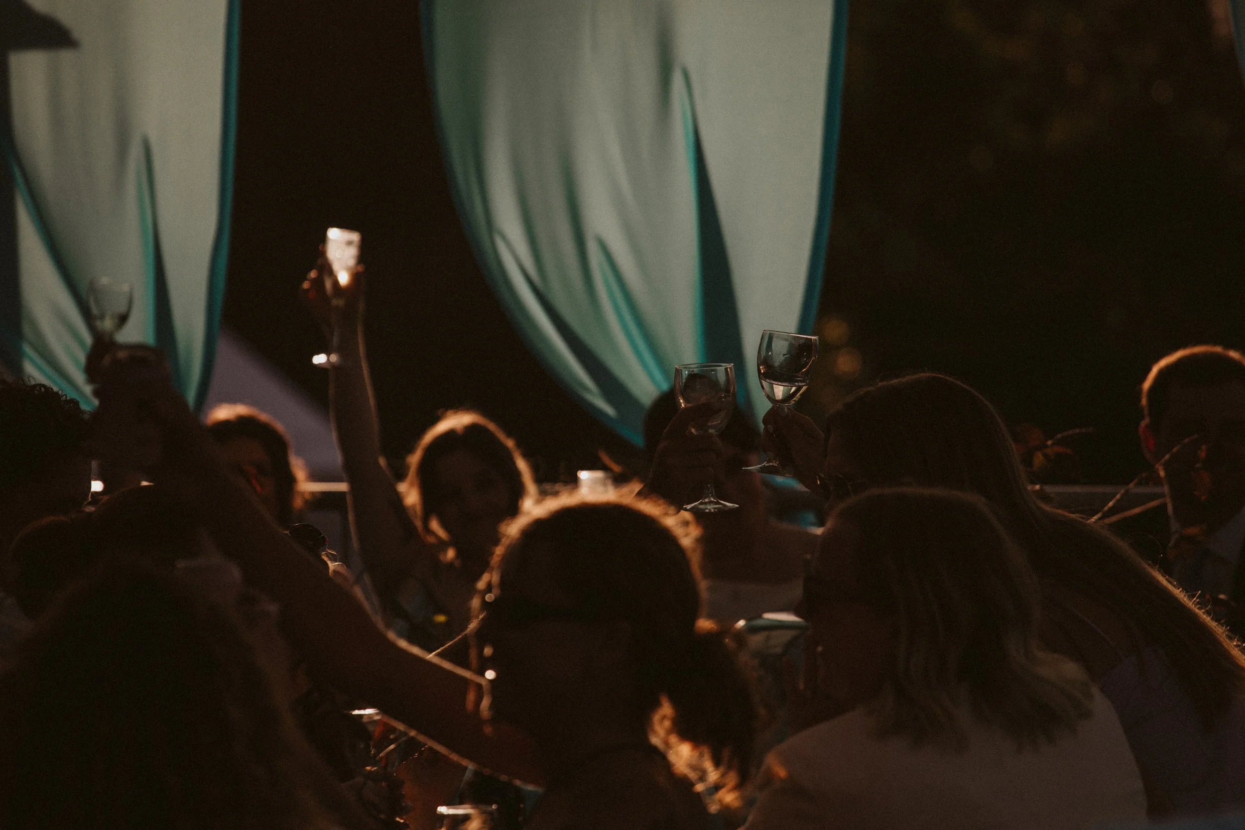 People at an outdoor event holding wine glasses, with large blue and white striped umbrellas in the background, during nighttime.