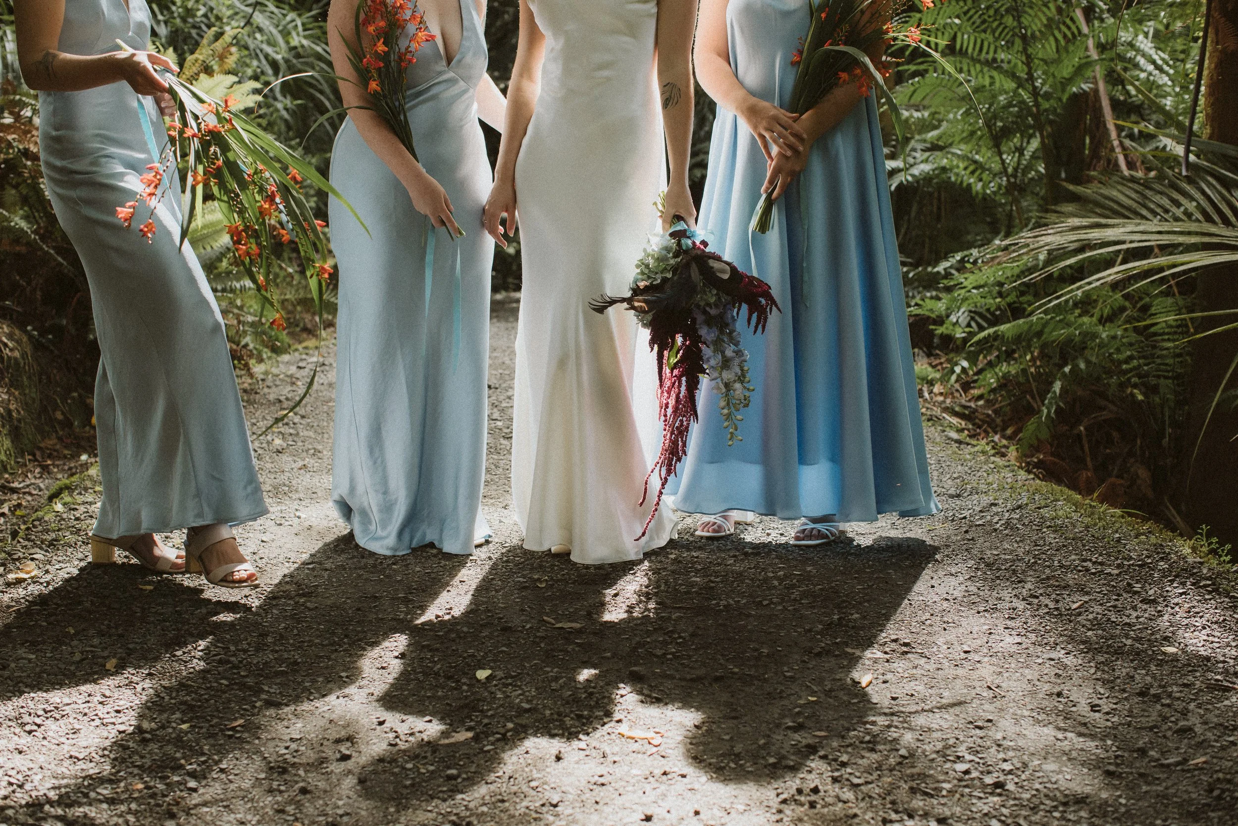 Women in dresses holding bouquets standing on a forest trail, with sunlight casting shadows on the ground.