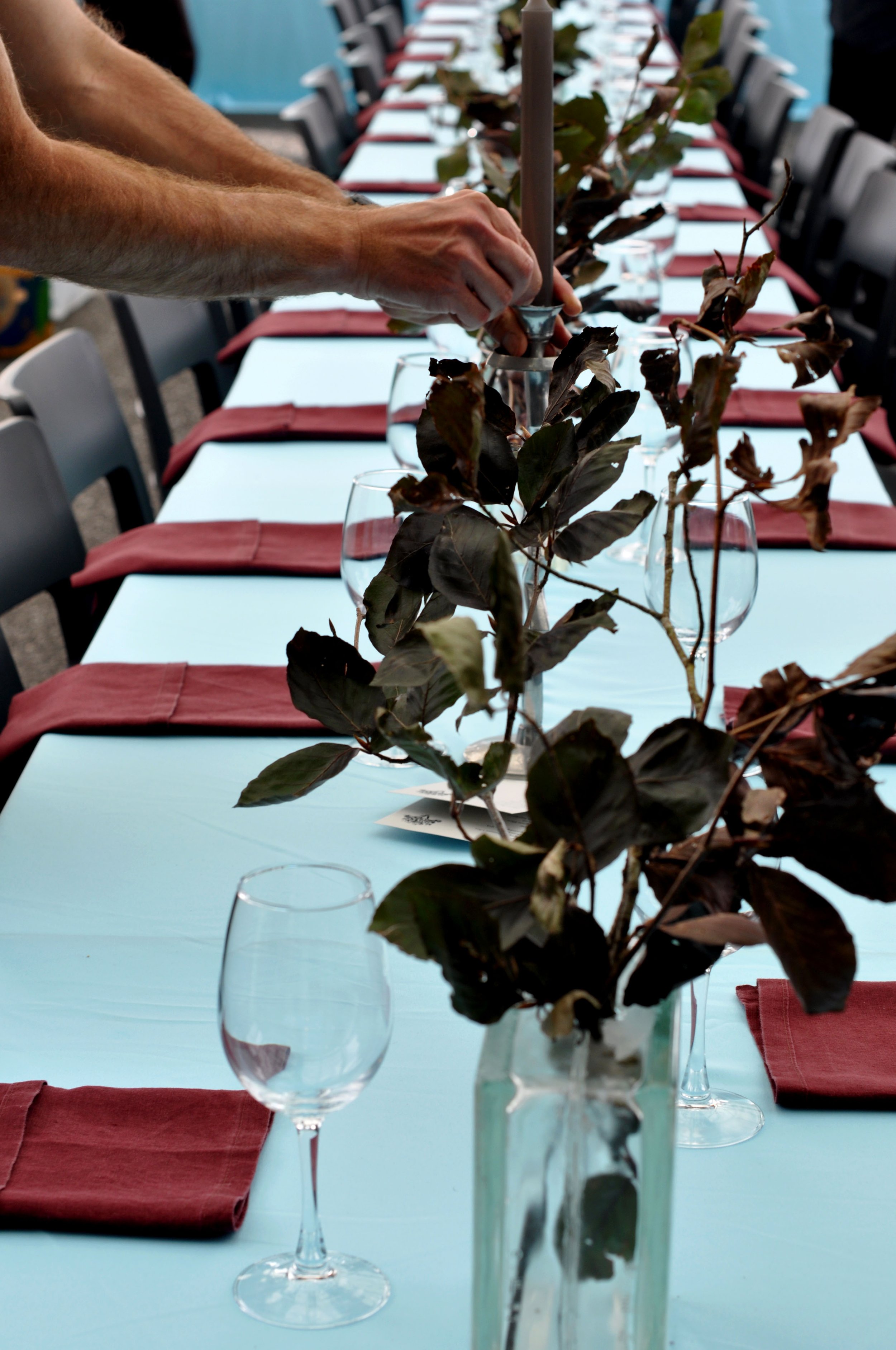 A long dining table decorated with a centerpiece of leafy branches in a tall glass vase, red napkins, and empty wine glasses, with chairs arranged on both sides.