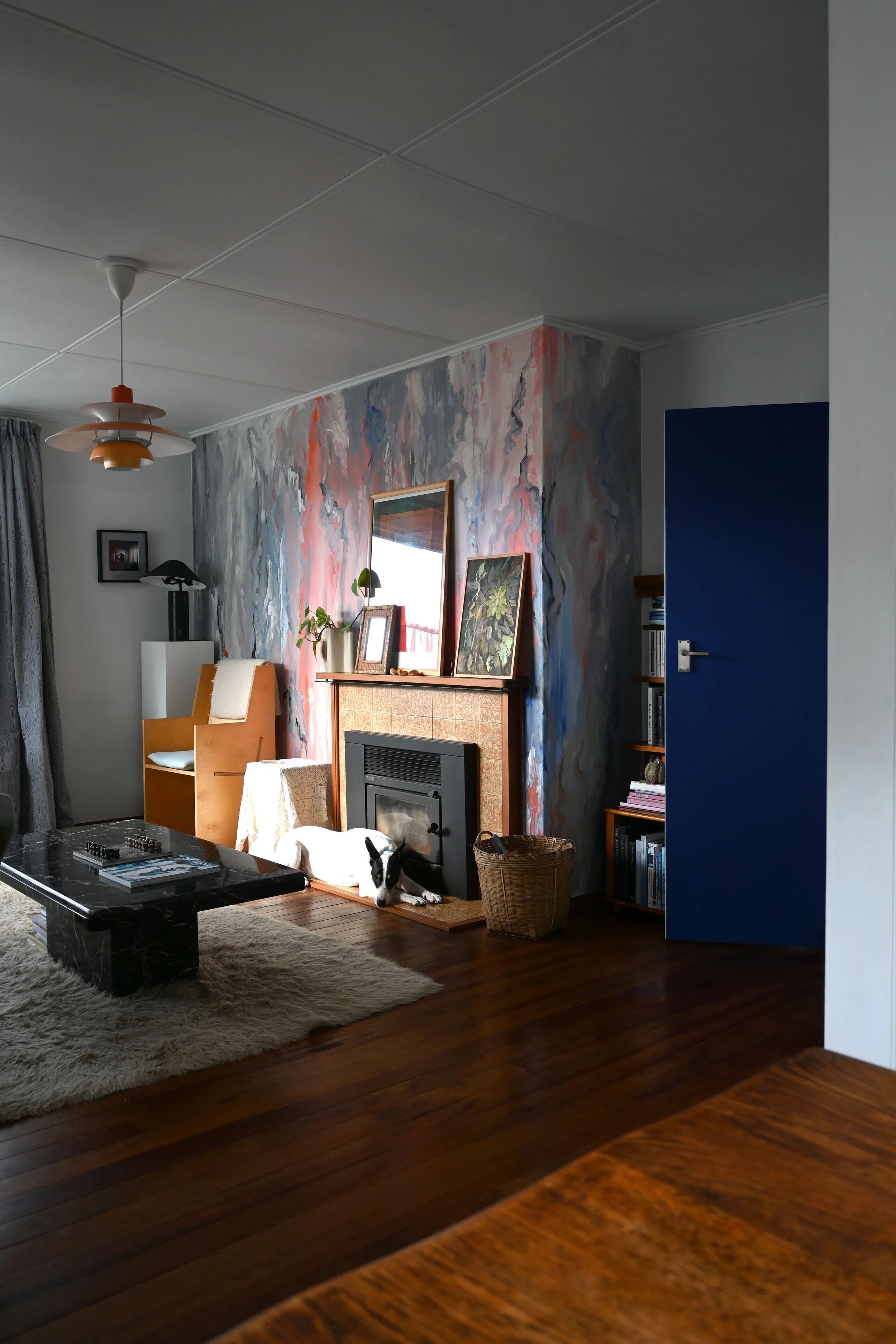 Interior of a living room with a blue door, abstract wall mural, a chimney with framed art on top, a black stove, a dog lying on a wooden floor, a beige rug, a black marble coffee table, a lamp, an armchair, and a window with curtains.