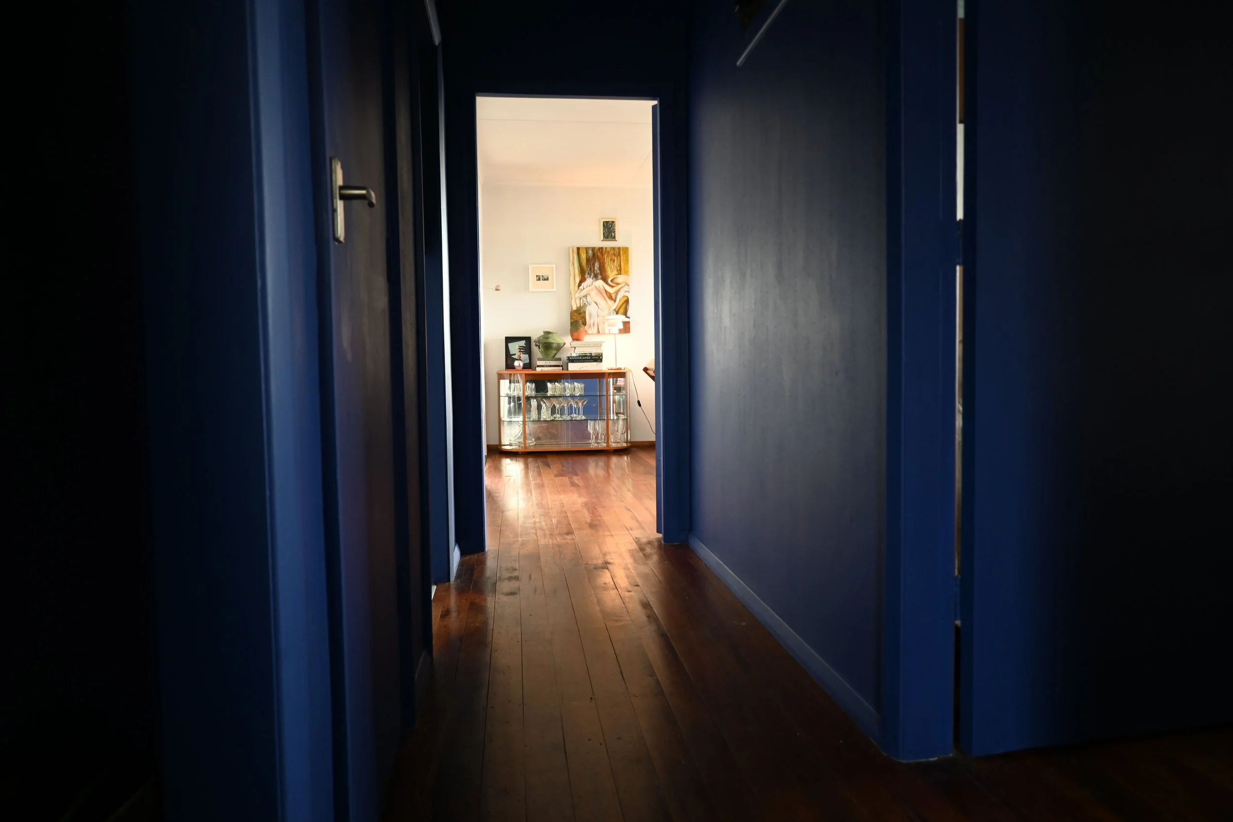 A view down a dimly lit hallway with blue walls and wooden flooring, leading to a sunlit room with artwork and a glass cabinet visible.