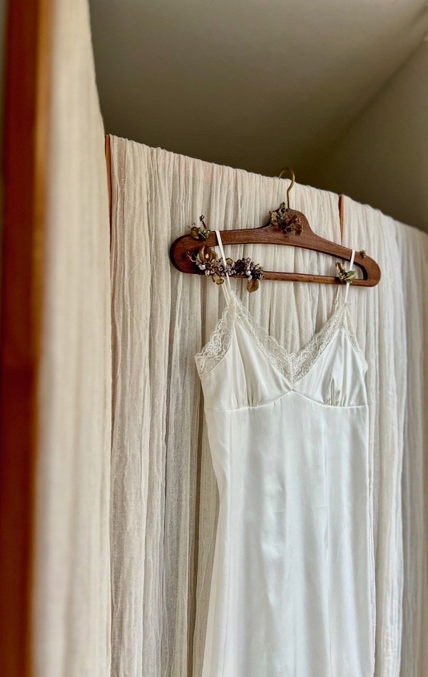 Wooden hanger decorated with beads and metal elements in a rustic style, holding a white lace slip dress against a neutral fabric background.