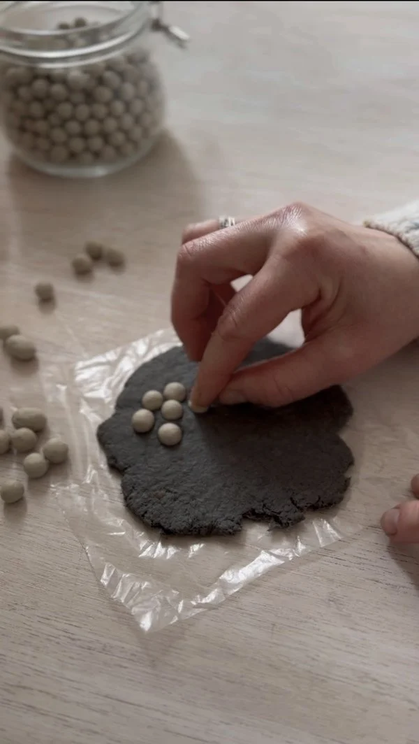A hand placing small ceramic baking beads onto rolled-out paper pulp, creating a unique texture. In the background, scattered beads and a jar filled with them are visible.