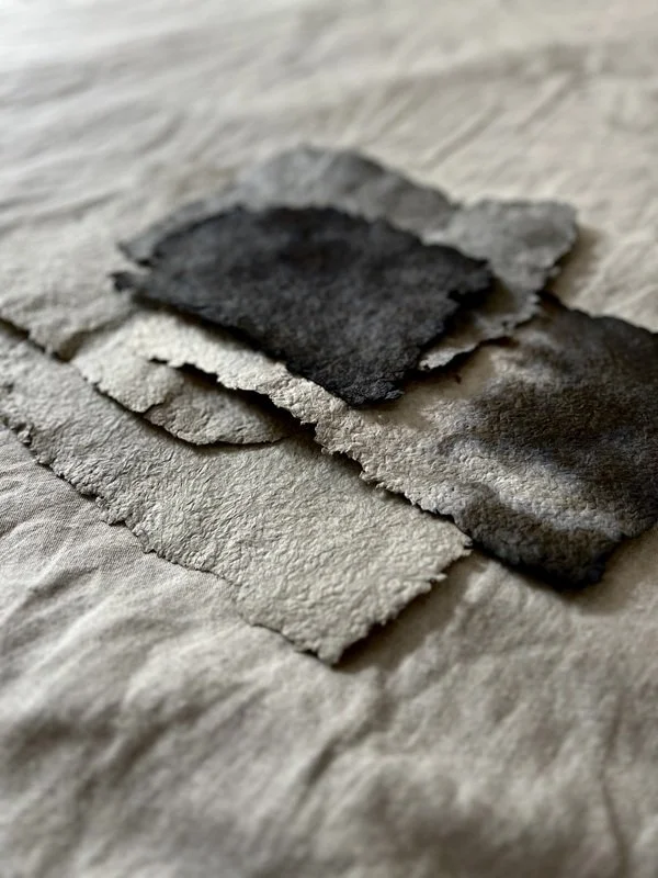 Close-up of handmade paper sheets with deckled edges in shades of gray and black, layered on a fabric surface. The texture of the paper and the soft, natural tones are highlighted, showing the unique handcrafted look