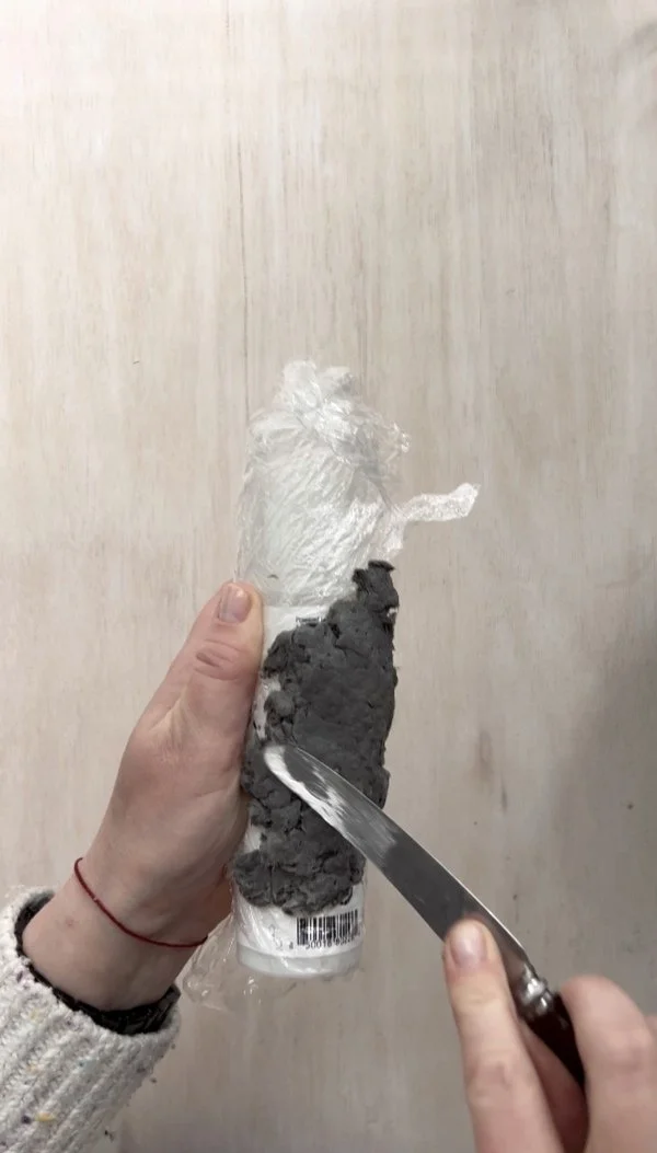 Hands using a knife to smooth paper pulp on a plastic-wrapped tube for creating a DIY napkin ring