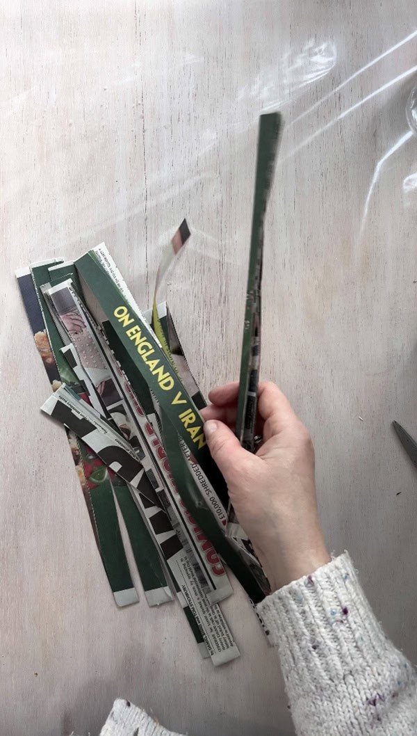 Hand holding strips of cut newspaper, with various colored pieces laid out on a light wooden surface. The strips are being prepared for use in a DIY handmade paper craft project.