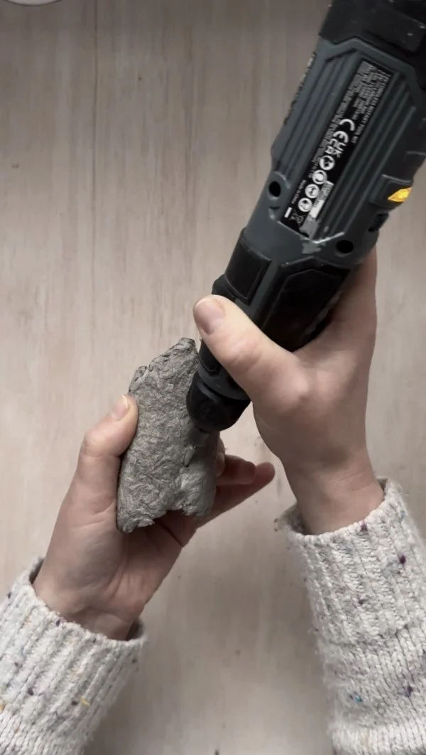 Hands using a drill to create holes in a dried paper pulp napkin ring as part of the DIY assembly process.