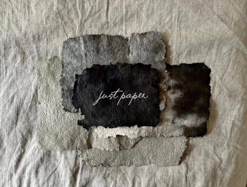 Various handmade paper sheets in different shades of gray and black, with the text 'just paper' written across the center. The paper sheets are placed on a neutral fabric background, showcasing their textures and natural tones.