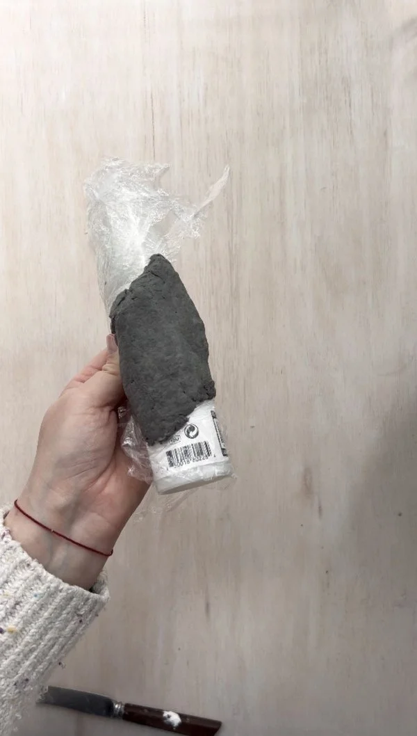 Hand holding a plastic-wrapped tube with a smooth layer of paper pulp applied to half of the surface, prepared to dry as part of a DIY napkin ring project.