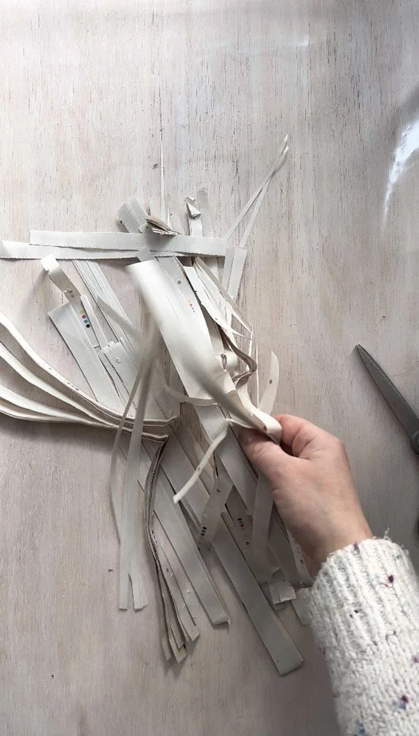 Hand holding strips of newspaper that have been cut into thin pieces, ready to be used in a handmade paper project. Scissors are visible on the side, with the strips arranged on a light wooden surface.