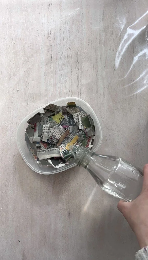 Water being poured from a bottle into a container filled with small newspaper strips to soak them for the handmade paper-making process.