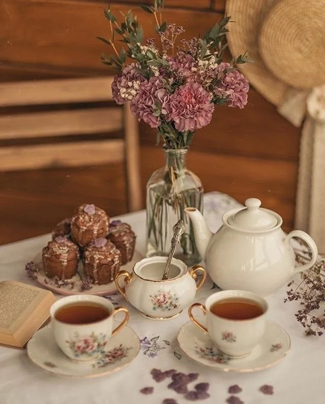 A floral arrangement of pink and purple flowers in a glass vase, two cups of tea, a teapot, a sugar bowl, a plate of cupcakes, an open book, and scattered petals on a table with a white tablecloth in a cozy wood-paneled room.