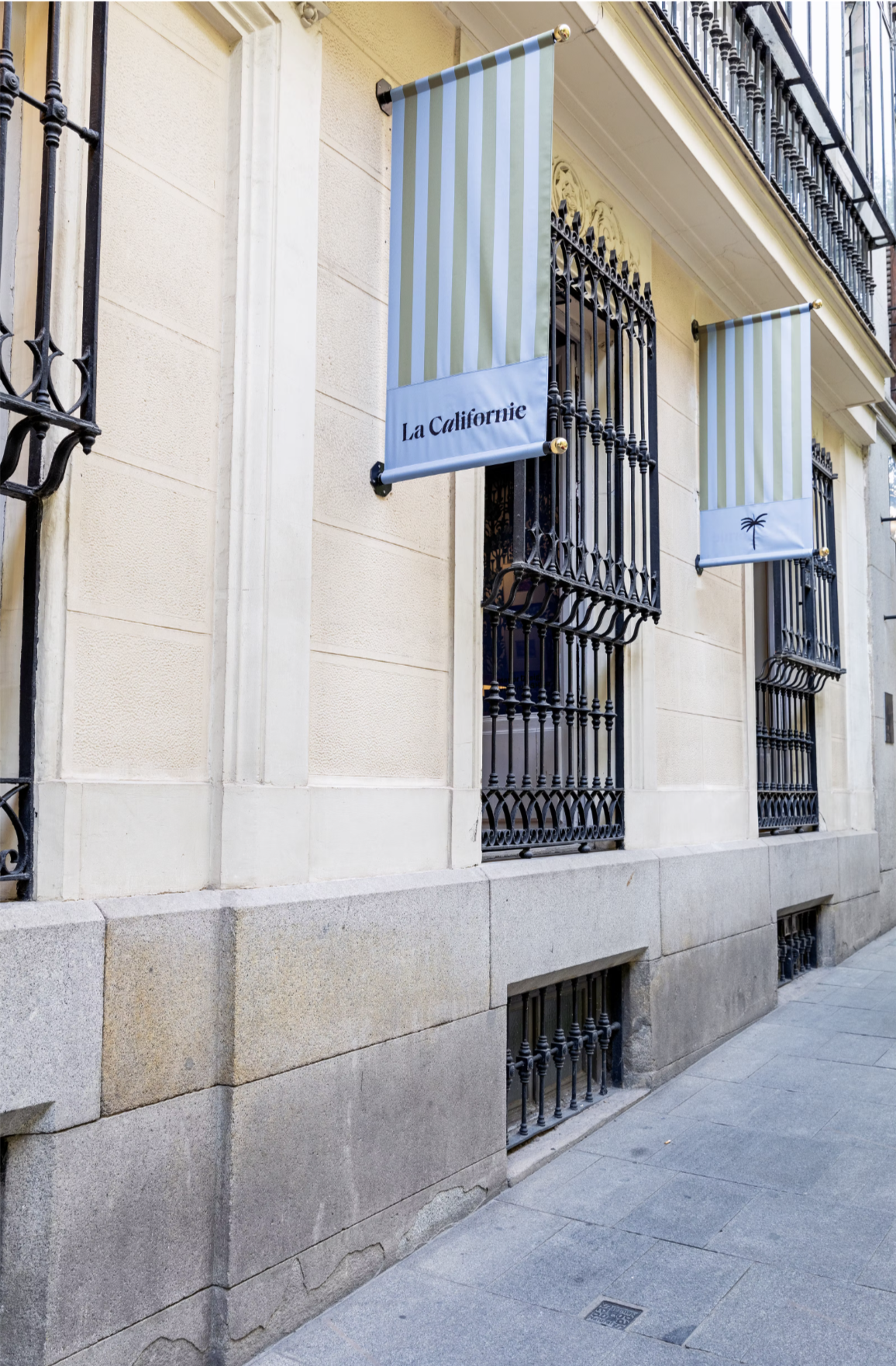 Two striped flags hanging on a building with ornate metal window grilles, one with the text 'La Californie' and the other with a palm tree icon, in an urban setting.