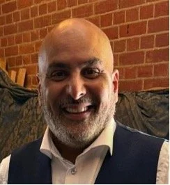 A smiling man with a bald head and gray beard, wearing a white shirt and dark vest, sitting in front of a brick wall.
