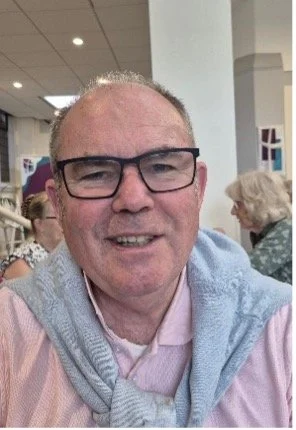 Close-up of a smiling man wearing glasses, a light pink shirt, and a gray sweater draped over his shoulders, in a social setting.