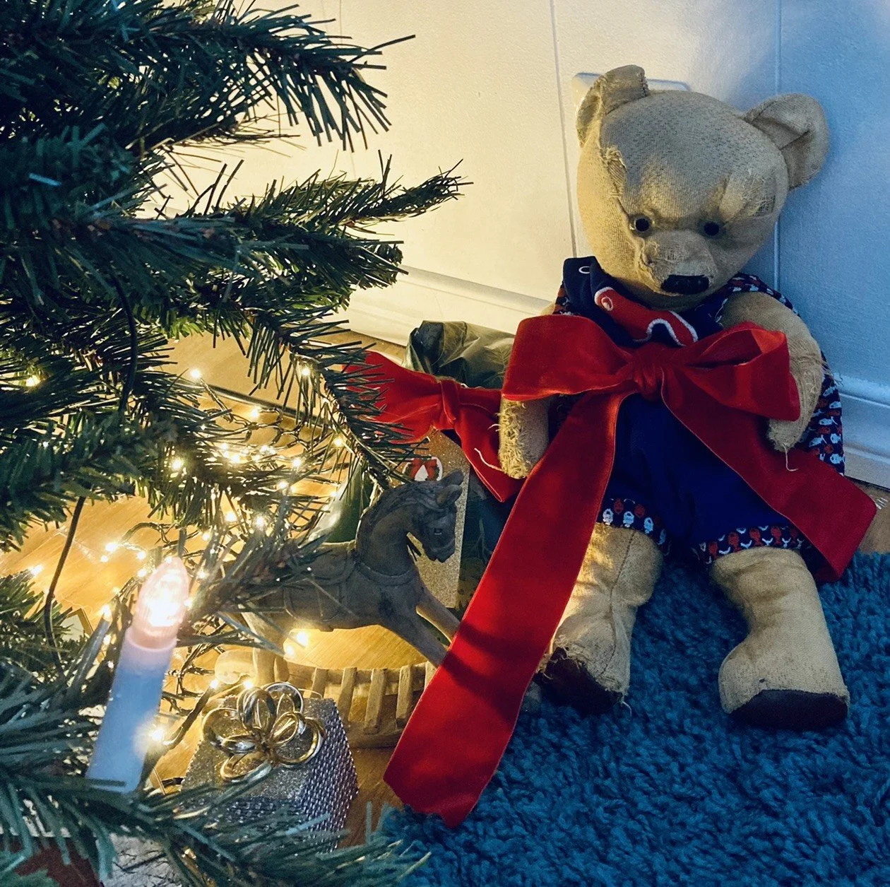 Bamse med rød sløyfe under juletre med lys, sammen med en liten gyngehest og en liten gave.