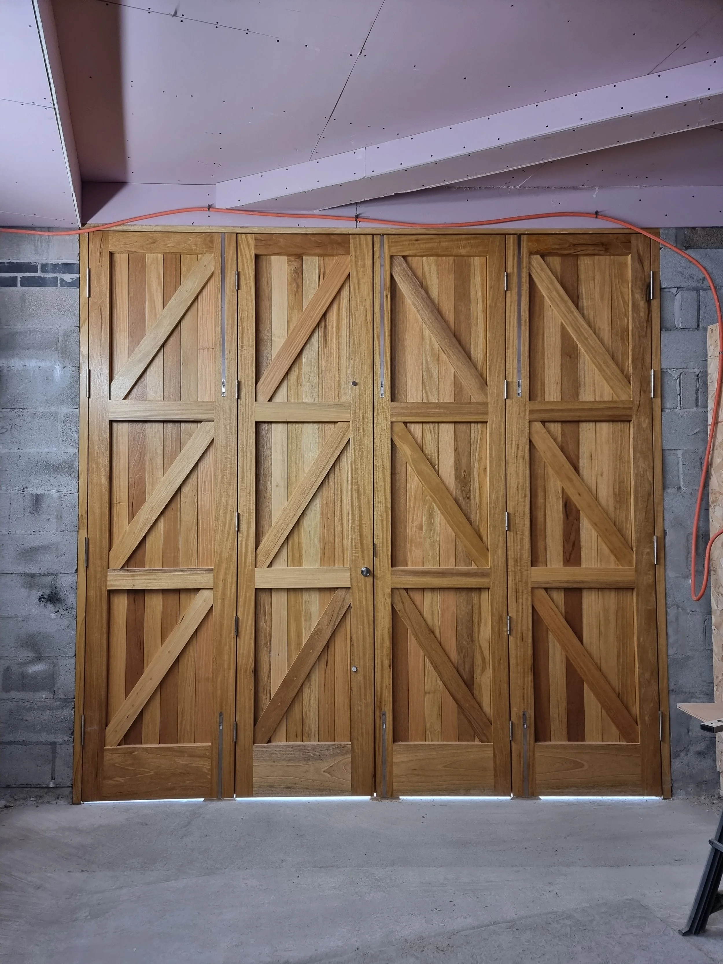 A set of four wooden barn doors in a construction area, with a concrete floor and bare concrete walls, partially opened at the bottom with a narrow gap showing outside light.