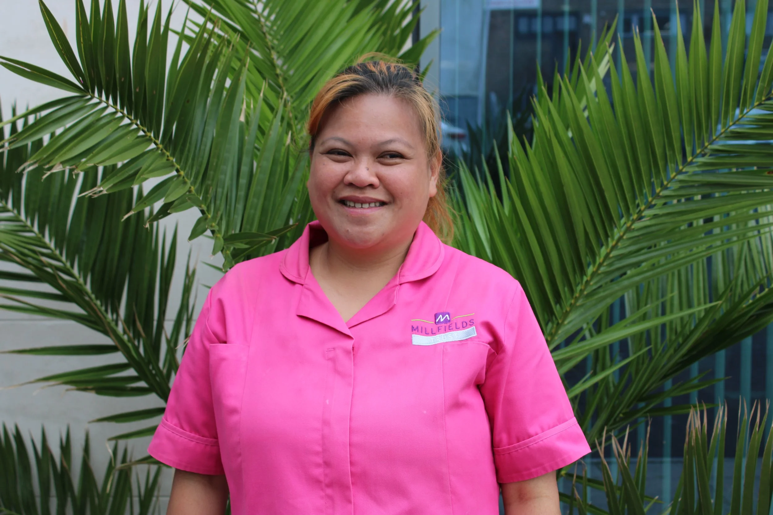 smiling woman, wearing bright pick uniform, stood in front of green plant with spiky leaves