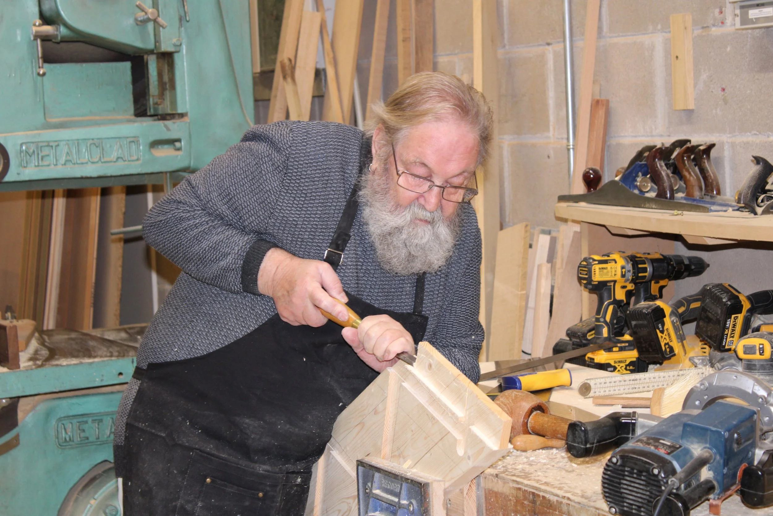 An elderly man with glasses and a beard working on a woodworking project in a workshop, using a chisel on a piece of wood.