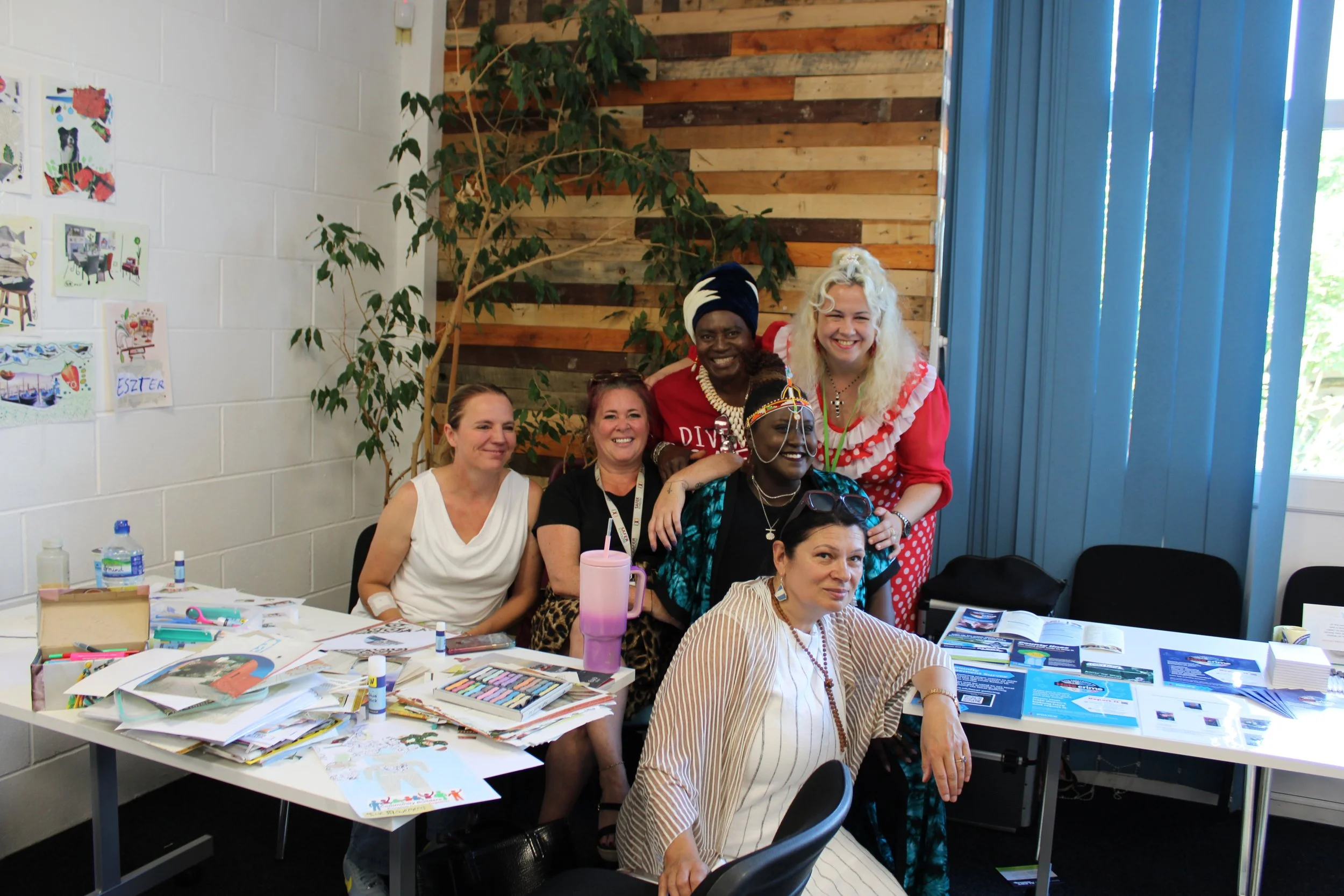 Group of six women smiling around a craft table in a room with a white brick wall, a wooden accent wall, and a window with blue curtains.