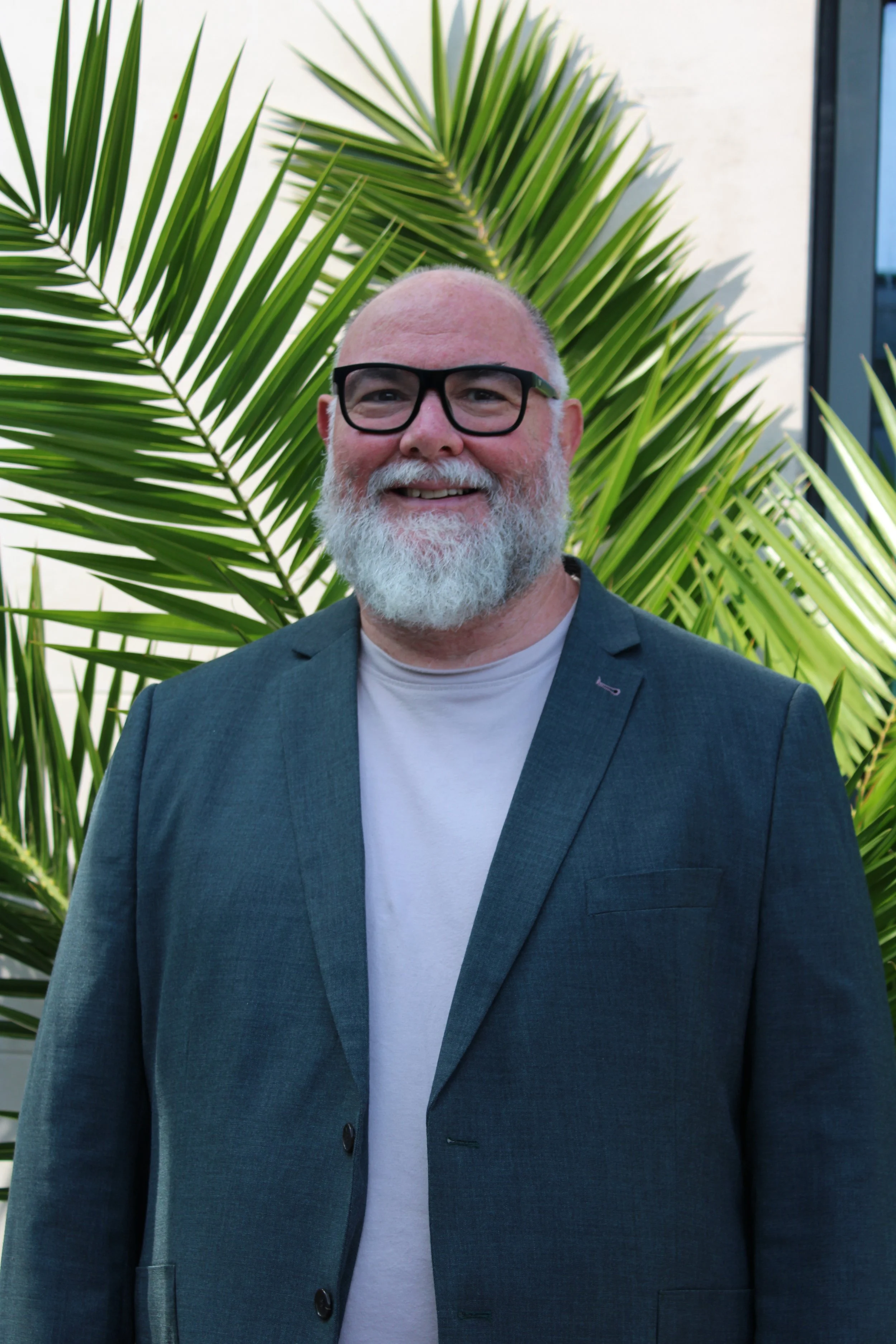 A smiling man with a white beard and glasses wearing a gray blazer and white t-shirt standing in front of large green palm leaves.