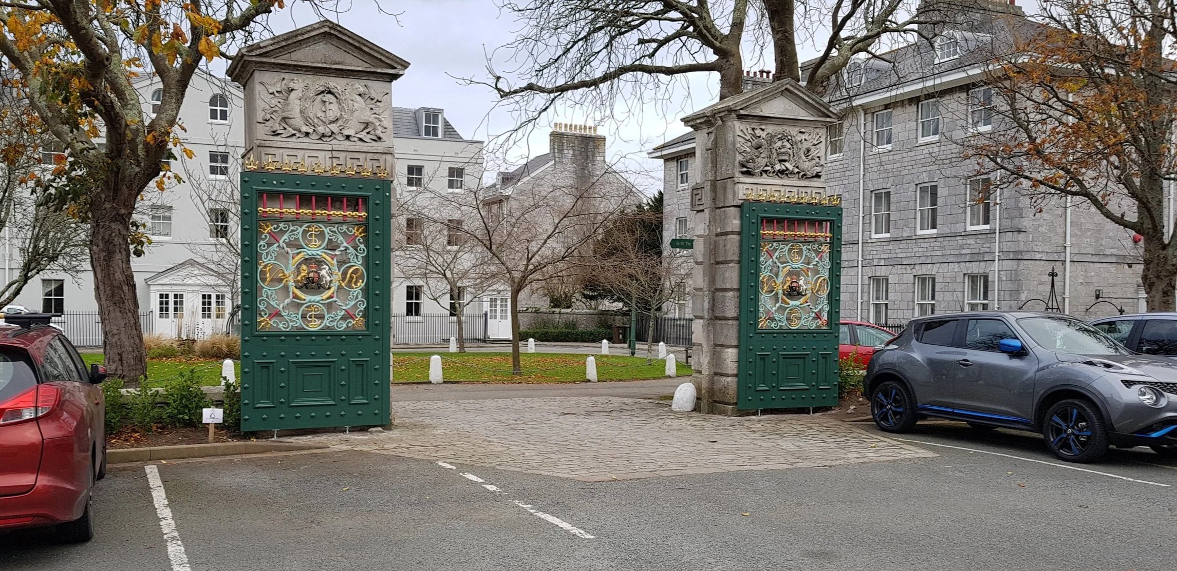 Parked cars next to ornate green metal gates with stone pillars, in front of residential buildings.