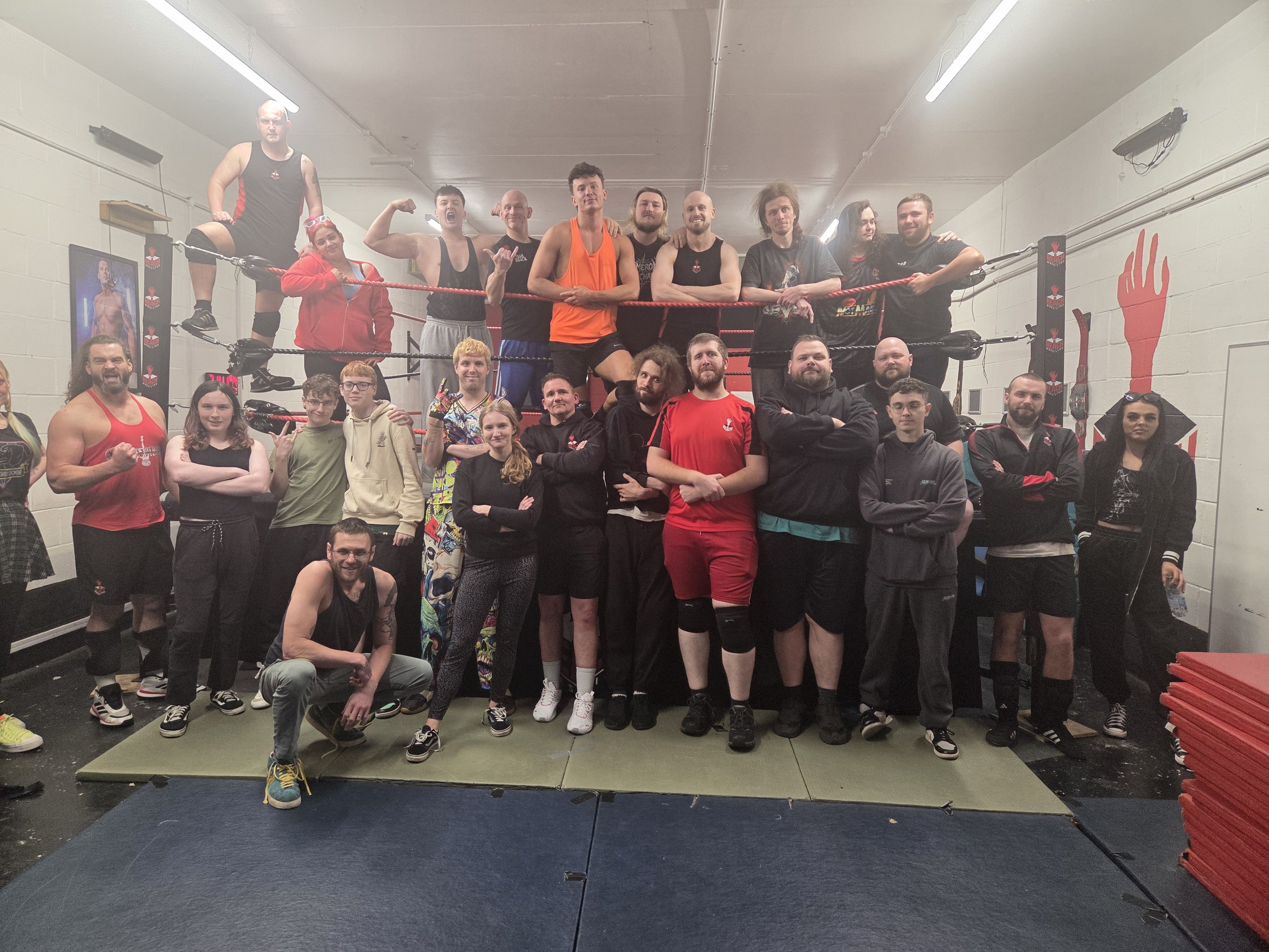 Group of people in a boxing gym posing for a photo around a boxing ring. Some are wearing athletic gear, and a few are making fists or flexing. The gym has posters and boxing equipment on the walls.
