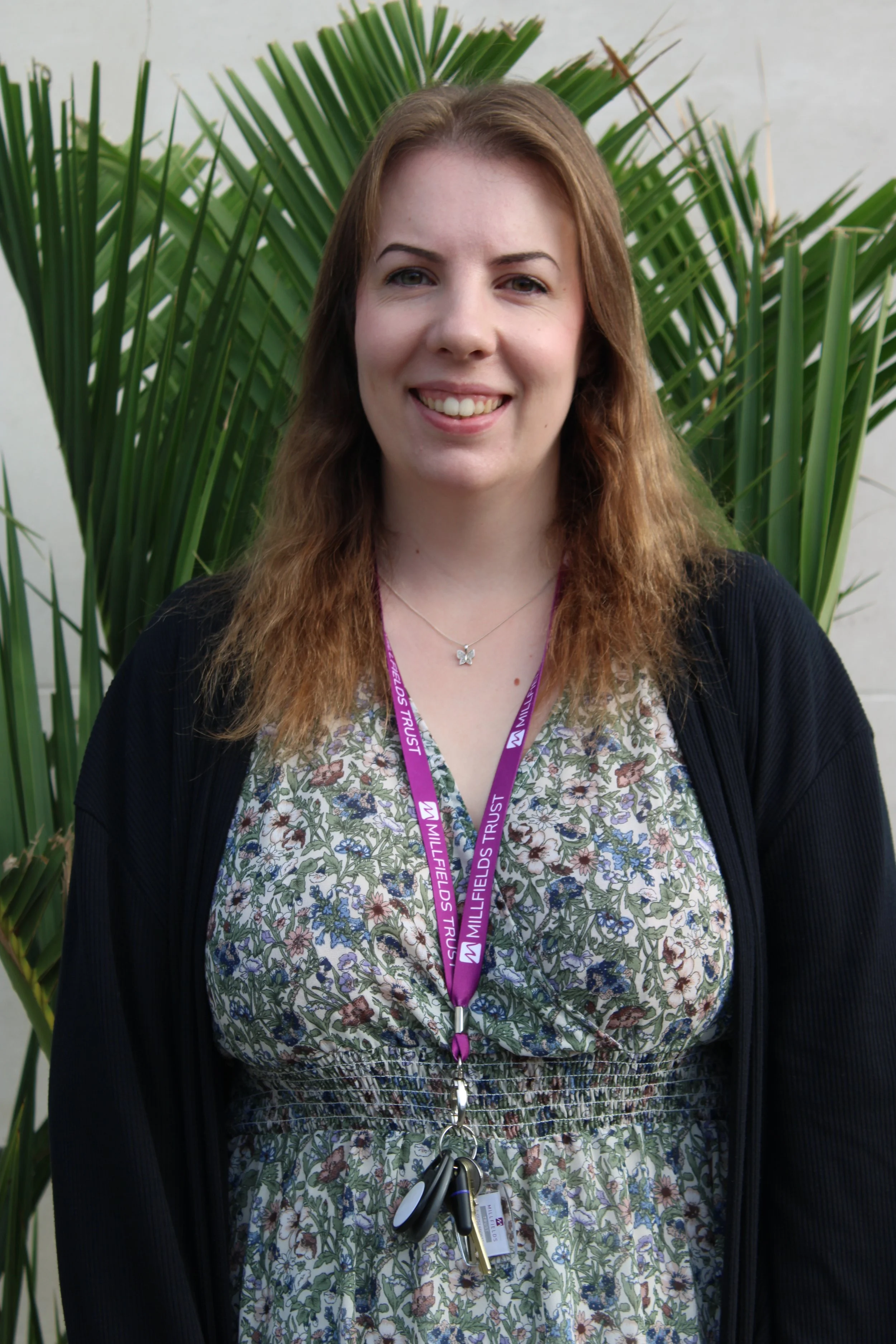 A woman with shoulder-length wavy brown hair, smiling, wearing a floral dress, a black cardigan, and a purple lanyard with white text, standing in front of a large green leafy plant.