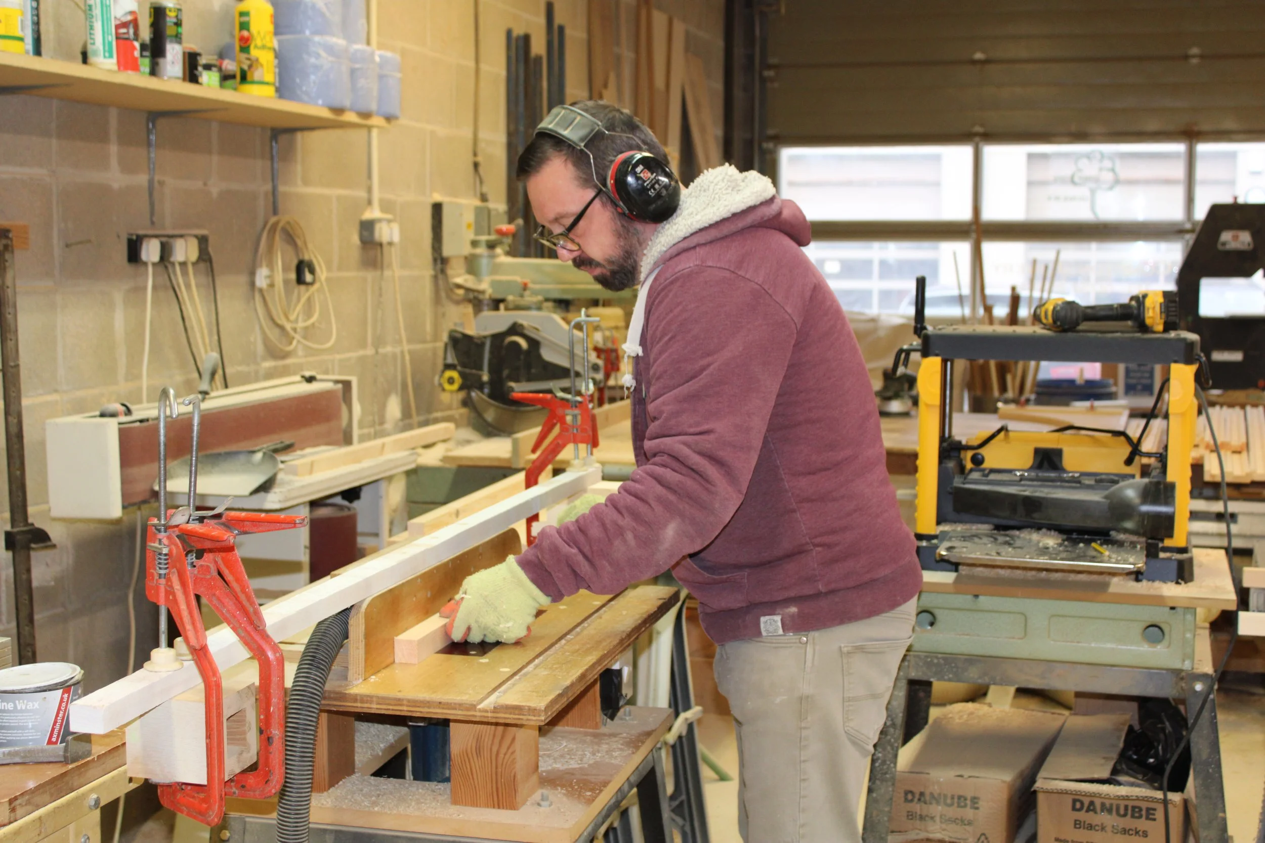 A man working in a woodworking shop, sanding or measuring a piece of wood while wearing ear protection and gloves, surrounded by woodworking tools and materials.