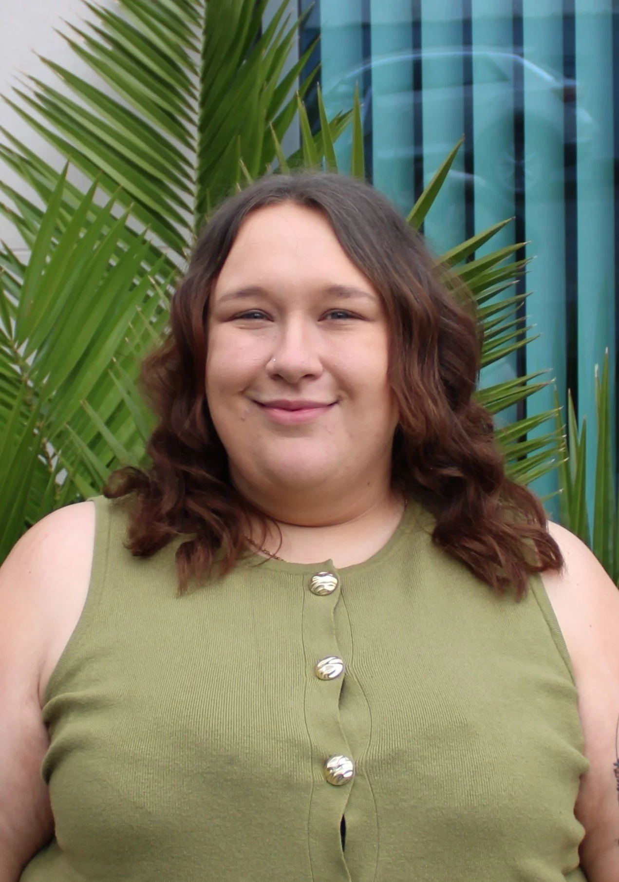 A smiling woman with wavy brown hair, wearing a green sleeveless top, standing in front of green palm leaves and a modern building with blue-tinted windows.