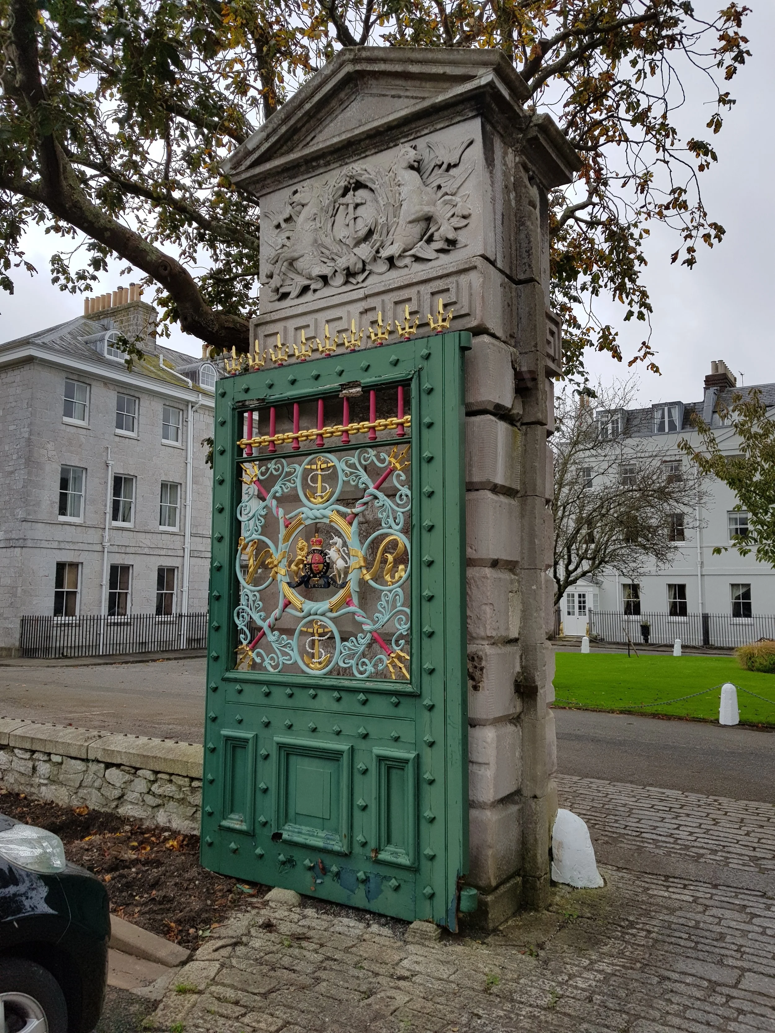 A historic stone gate brick wall with an ornate iron gate painted green and decorated with gold accents. The stone pillar above the gate features a carved relief of a dragon and other figures. In the background, there are large white multi-story houses and a large tree with some leaves falling.