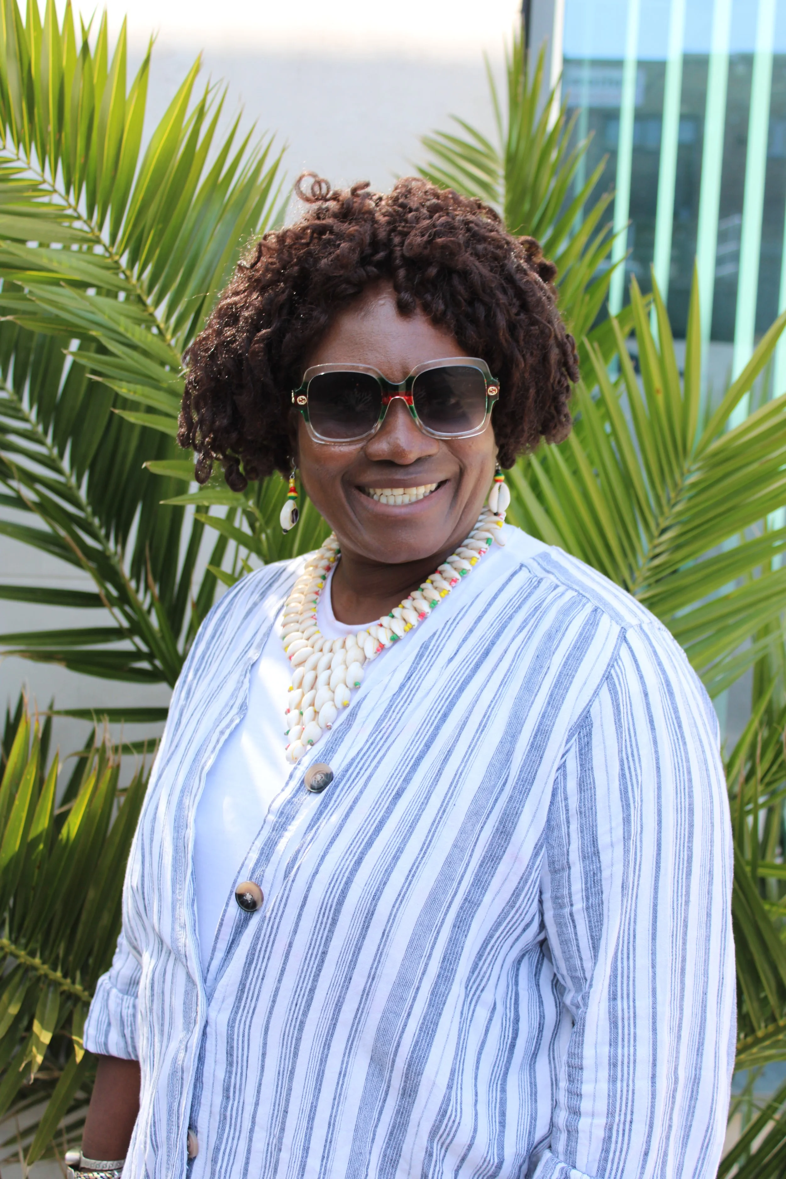 A woman with dark skin, curly short hair, and sunglasses smiling at the camera, standing in front of green palm leaves and modern glass buildings.