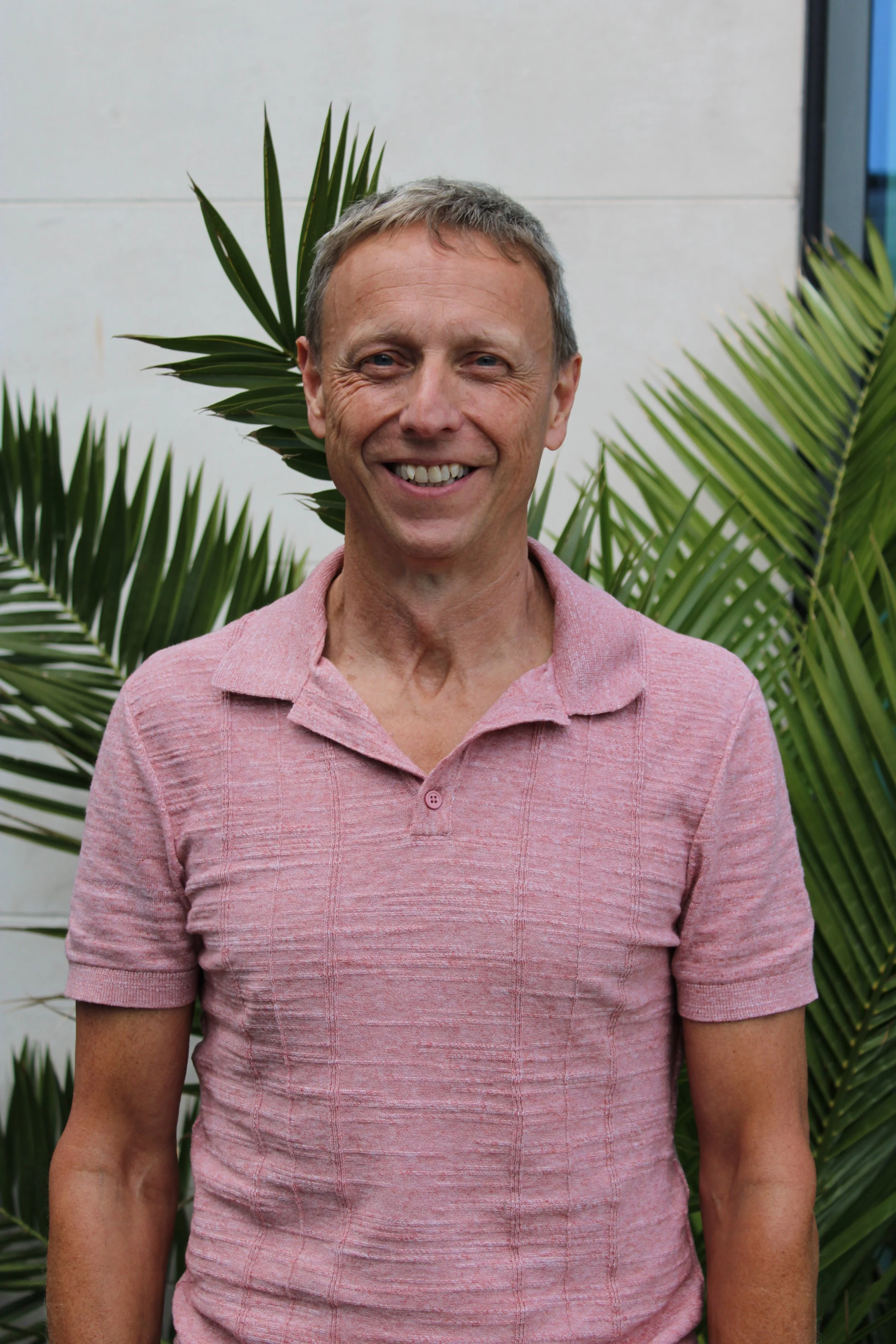 A man with short, gray hair and blue eyes smiling, wearing a pink polo shirt, standing outdoors in front of green palm plants and a white wall.