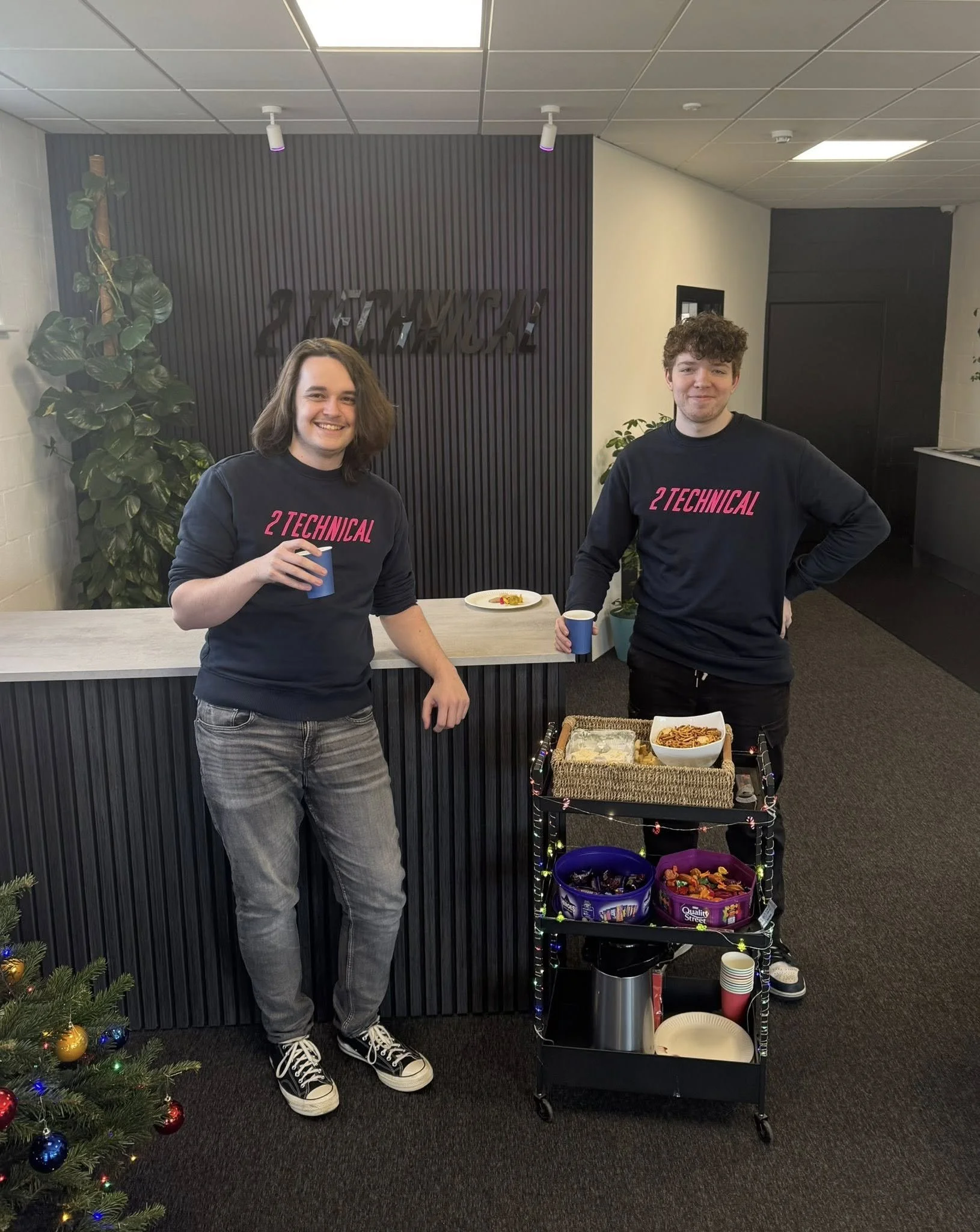 Two young men standing at a reception desk, holding blue cups, wearing matching black sweatshirts with pink text that reads '2 TECHNICAL.' They are smiling, with a snack cart filled with chips, candies, and cups nearby. A small decorated Christmas tree is visible in the lower left corner.