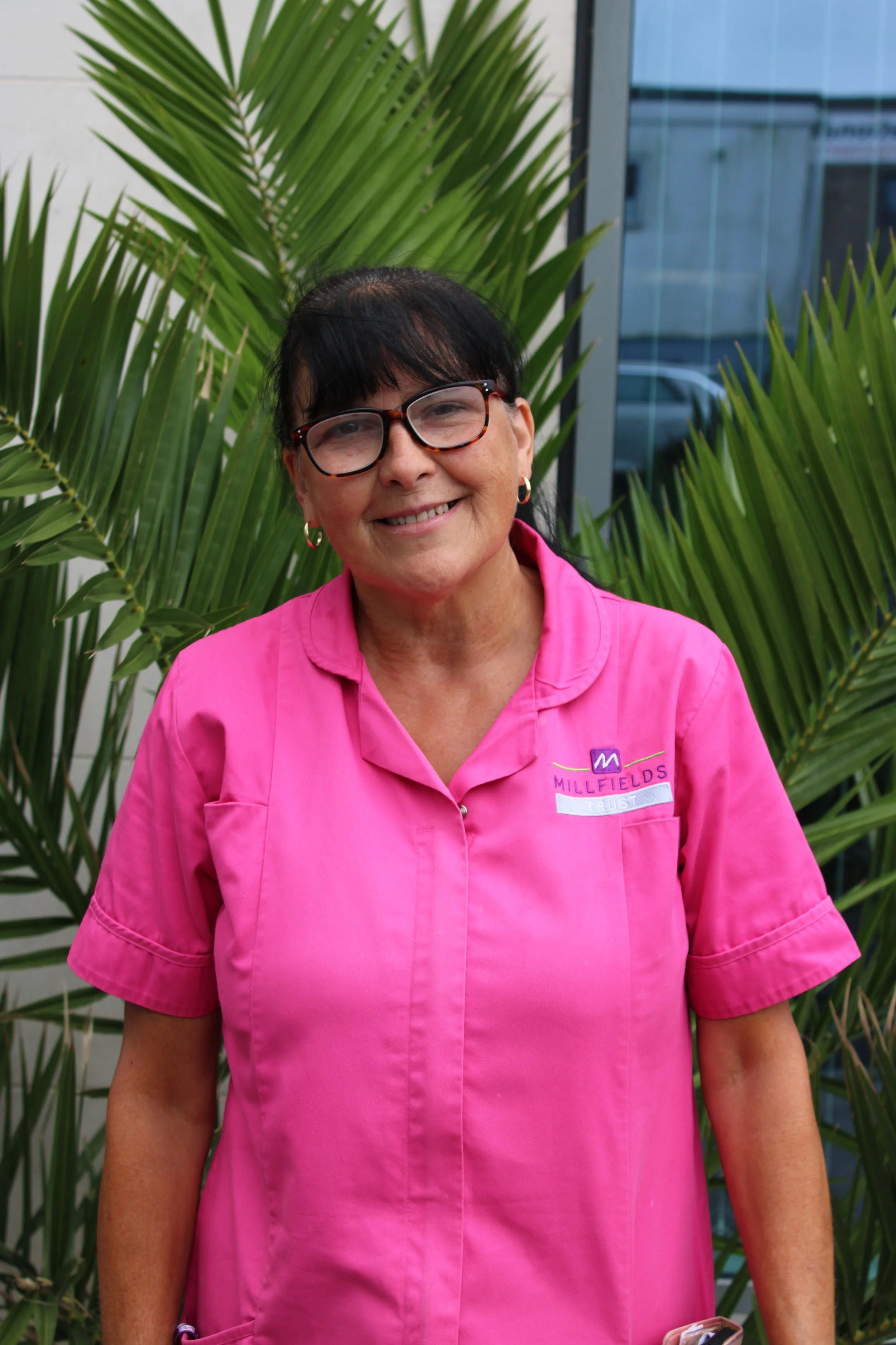 A woman with dark hair, wearing glasses, in a bright pink uniform, stands outdoors in front of a large plant with pointed leaves, with a light-colored wall in the background.