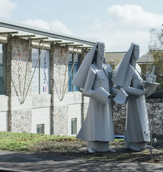 Two large gray statues of stylized women/nurses holding objects, located outside a modern building.