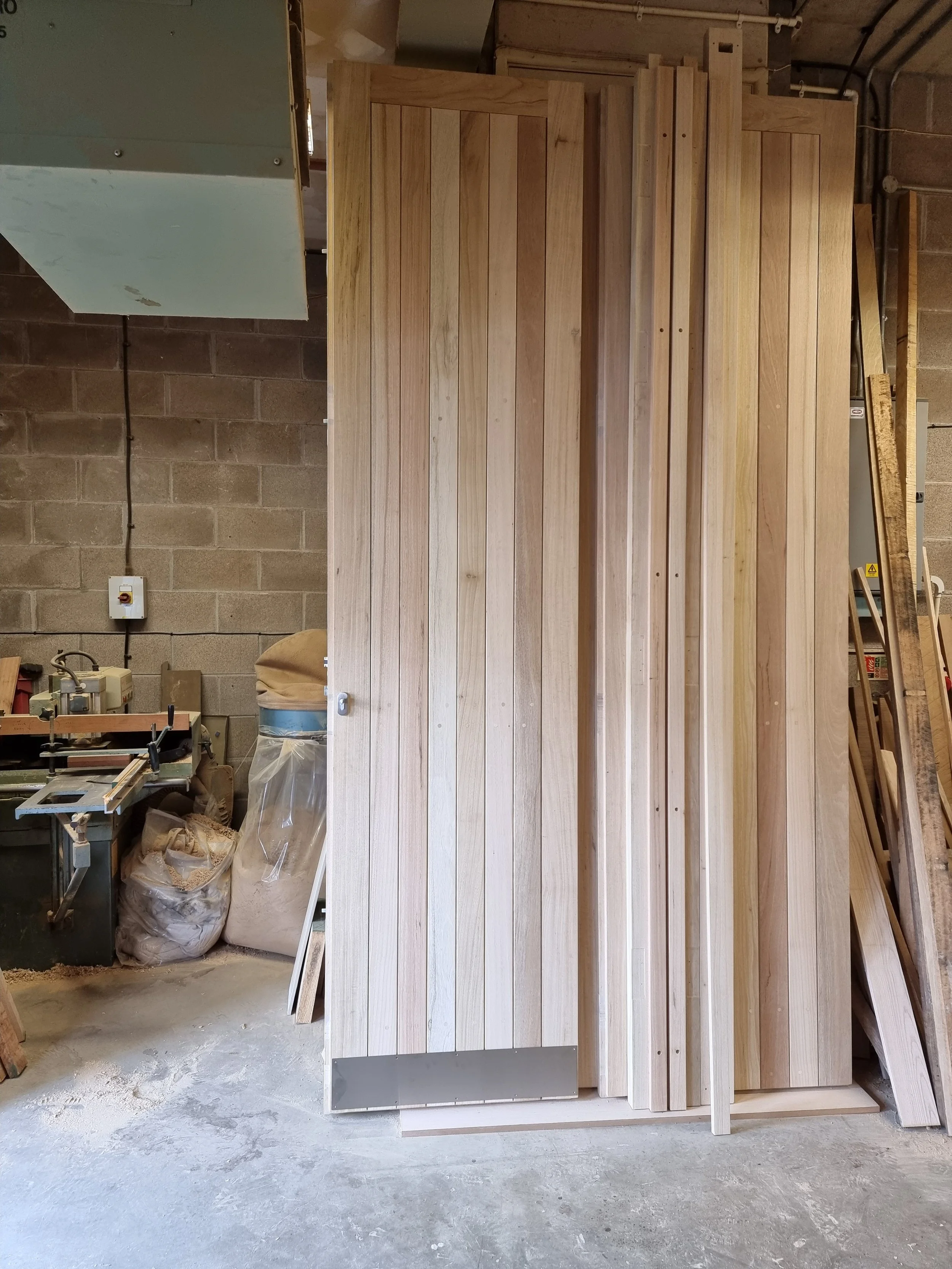 Stack of unfinished wooden planks stored vertically in a workshop with a concrete floor, brick wall, and woodworking tools in the background.