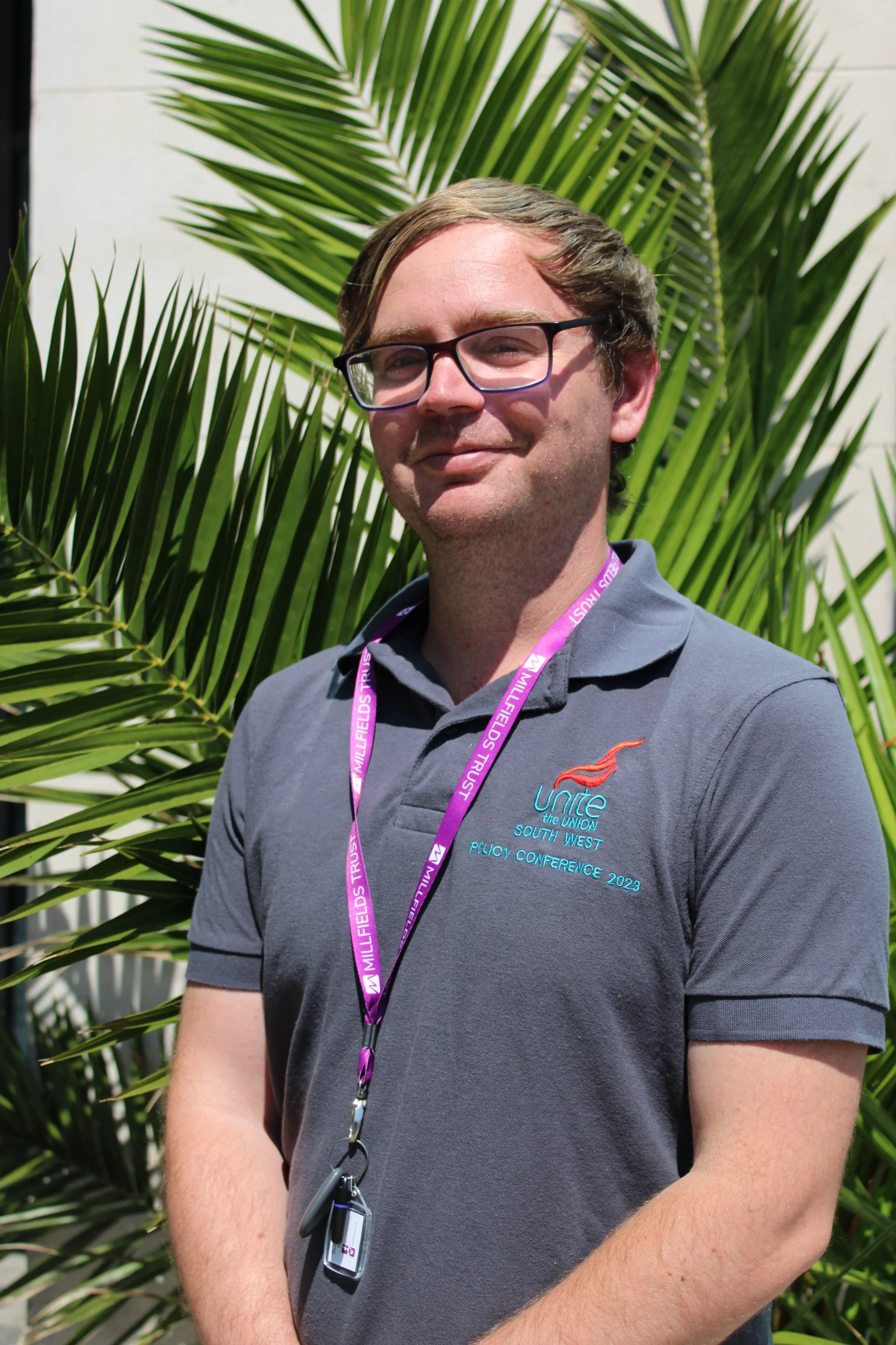 A man with glasses wearing a gray polo shirt standing outdoors in front of green palm leaves, with a conference badge around his neck.