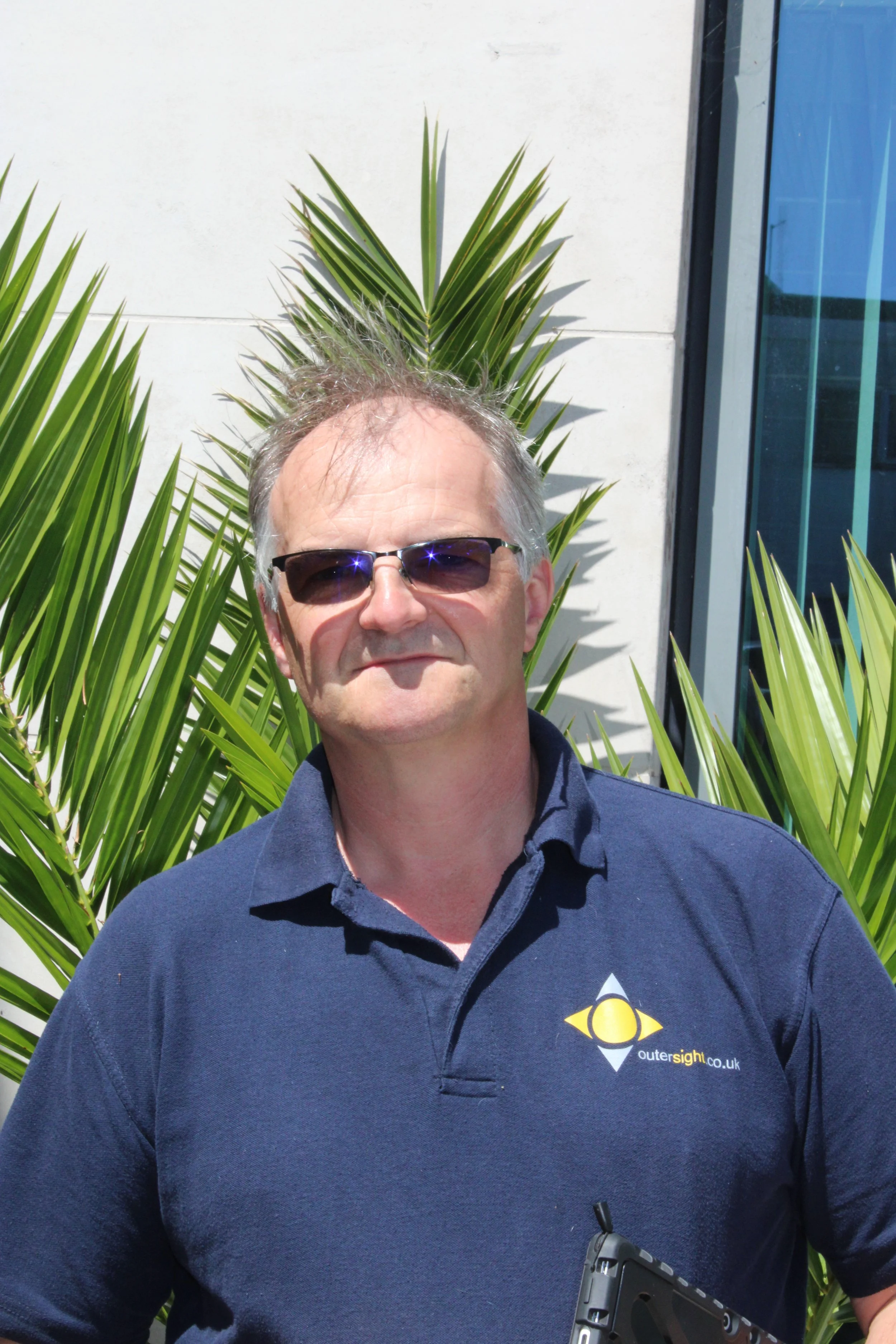 A man with gray hair and sunglasses standing outdoors in front of a white wall and green palm leaves, wearing a navy blue polo shirt with a logo that reads 'outer sight.co.uk'.