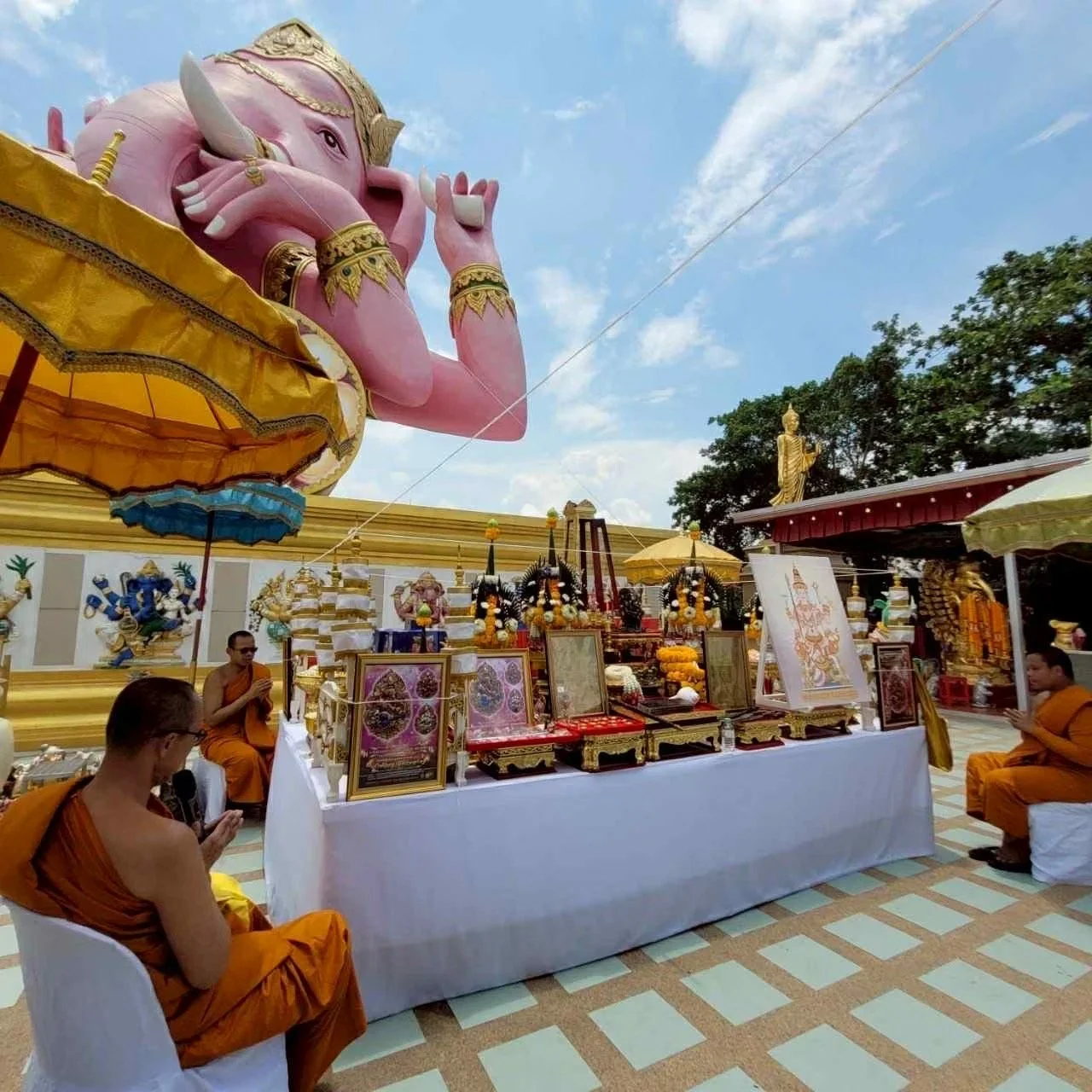 Large pink Ganesha statue with gold accents, surrounded by offerings, monks, and religious images during a ceremony at a temple.