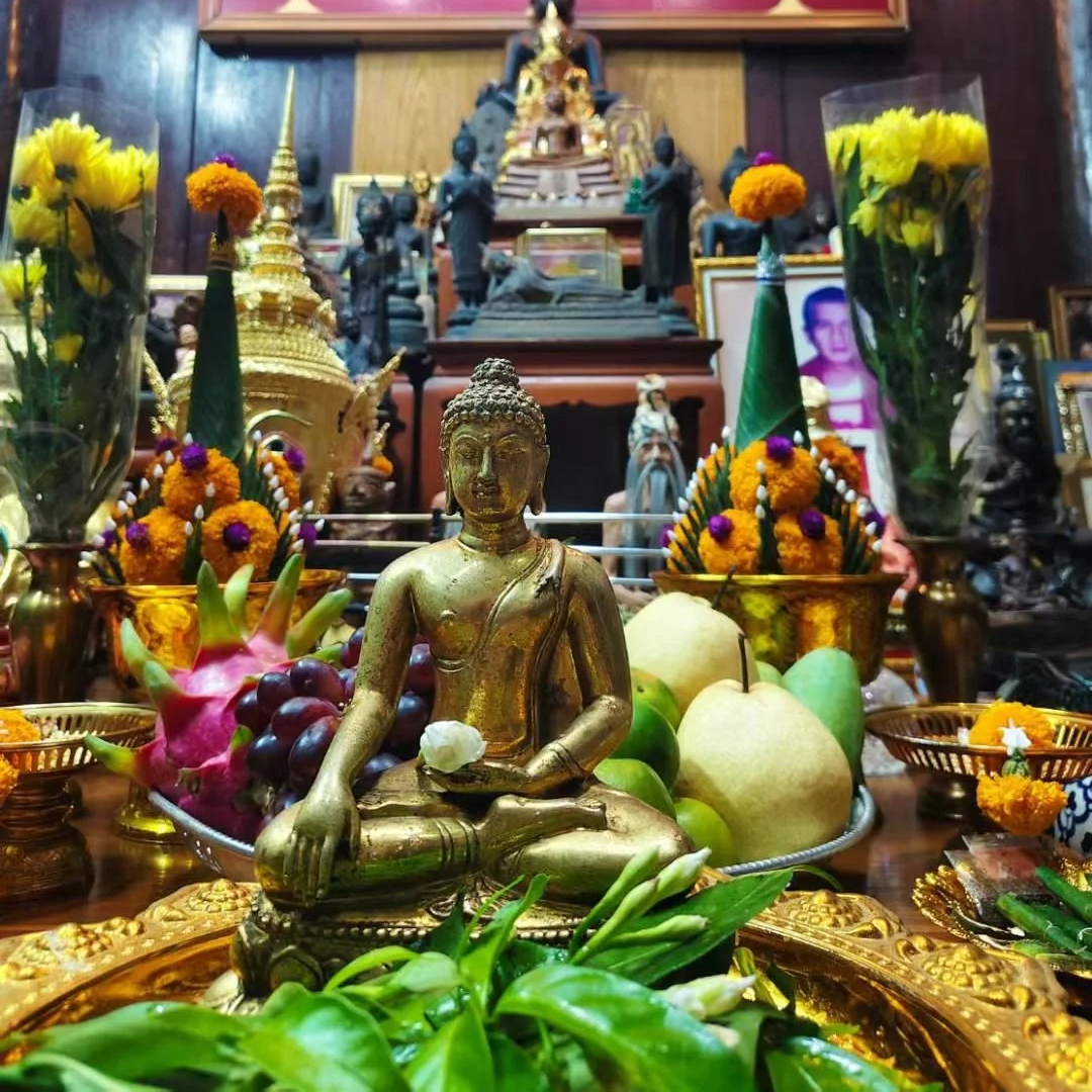 A golden Buddha statue seated on a decorative gold tray surrounded by fresh fruits and flowers, with an elaborate altar in the background filled with smaller statues, framed photos, and offerings.