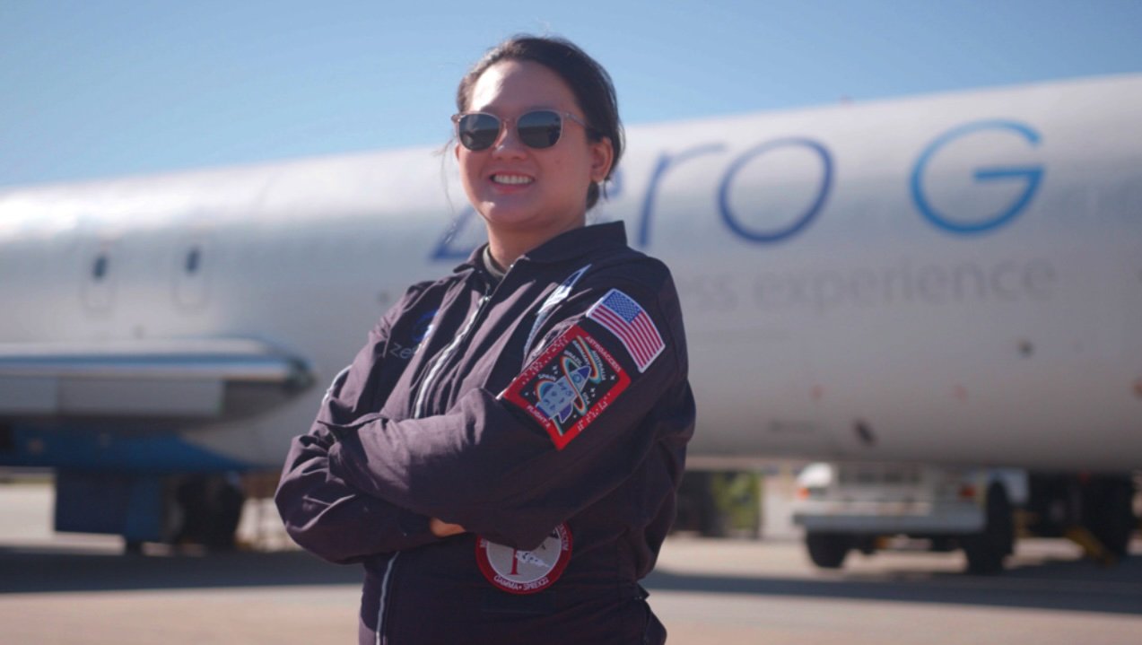 Sheila poses next to the Zero Gravity plane in an AstroAccess flight suit. Her arms are crossed and she wears aviator sunglasses while smiling at the camera.