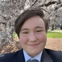 Photo of Emmett Lockwood, a white passing mixed white and First Nations individual with brown hair wearing a blue suit jacket, blue collared shirt and grey tie smiling while standing infront of a cherry blossom bush.